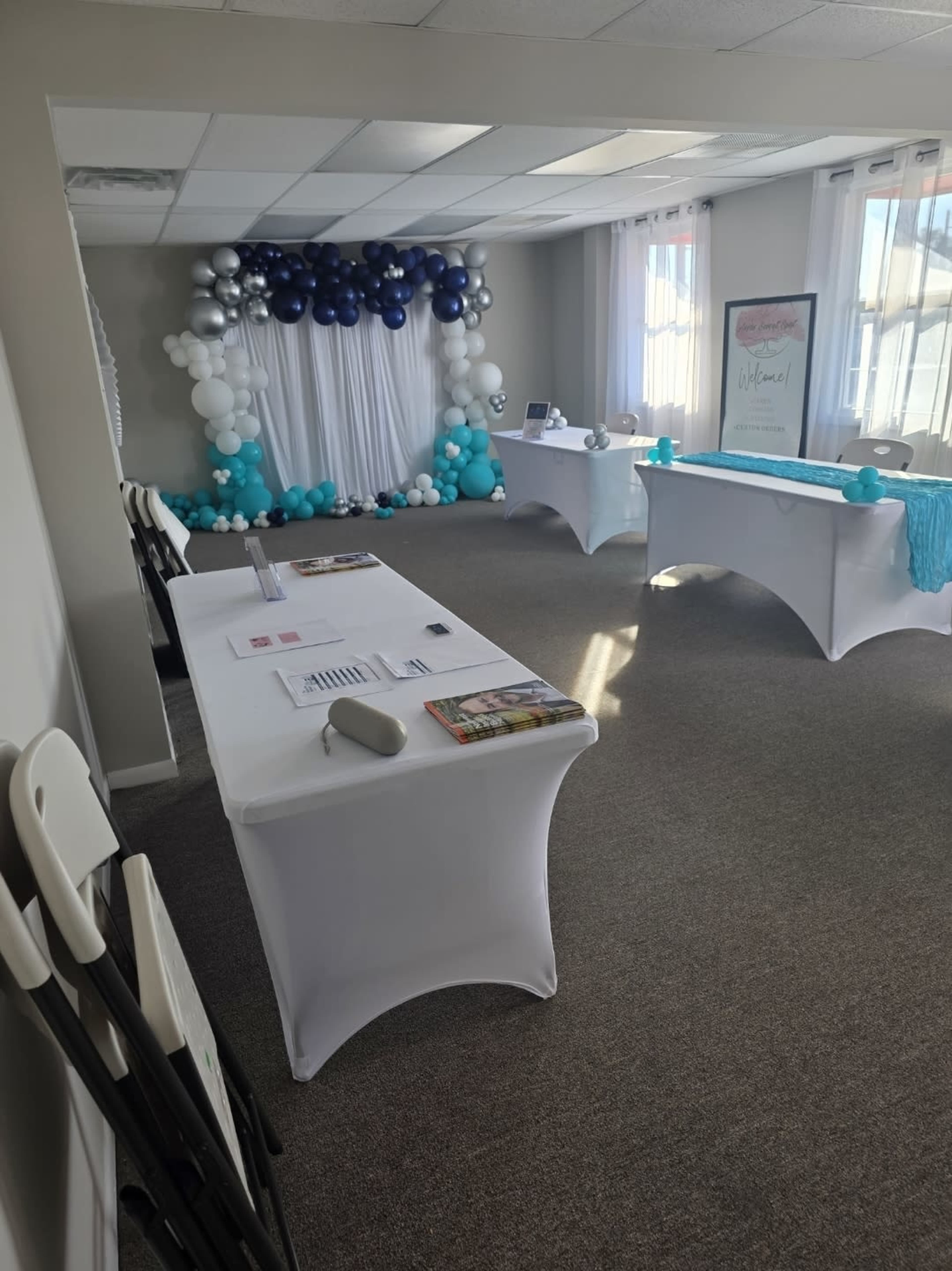 The image shows an event space decorated with a blue and white balloon arch, white tables covered in tablecloths, and promotional materials displayed on the tables.