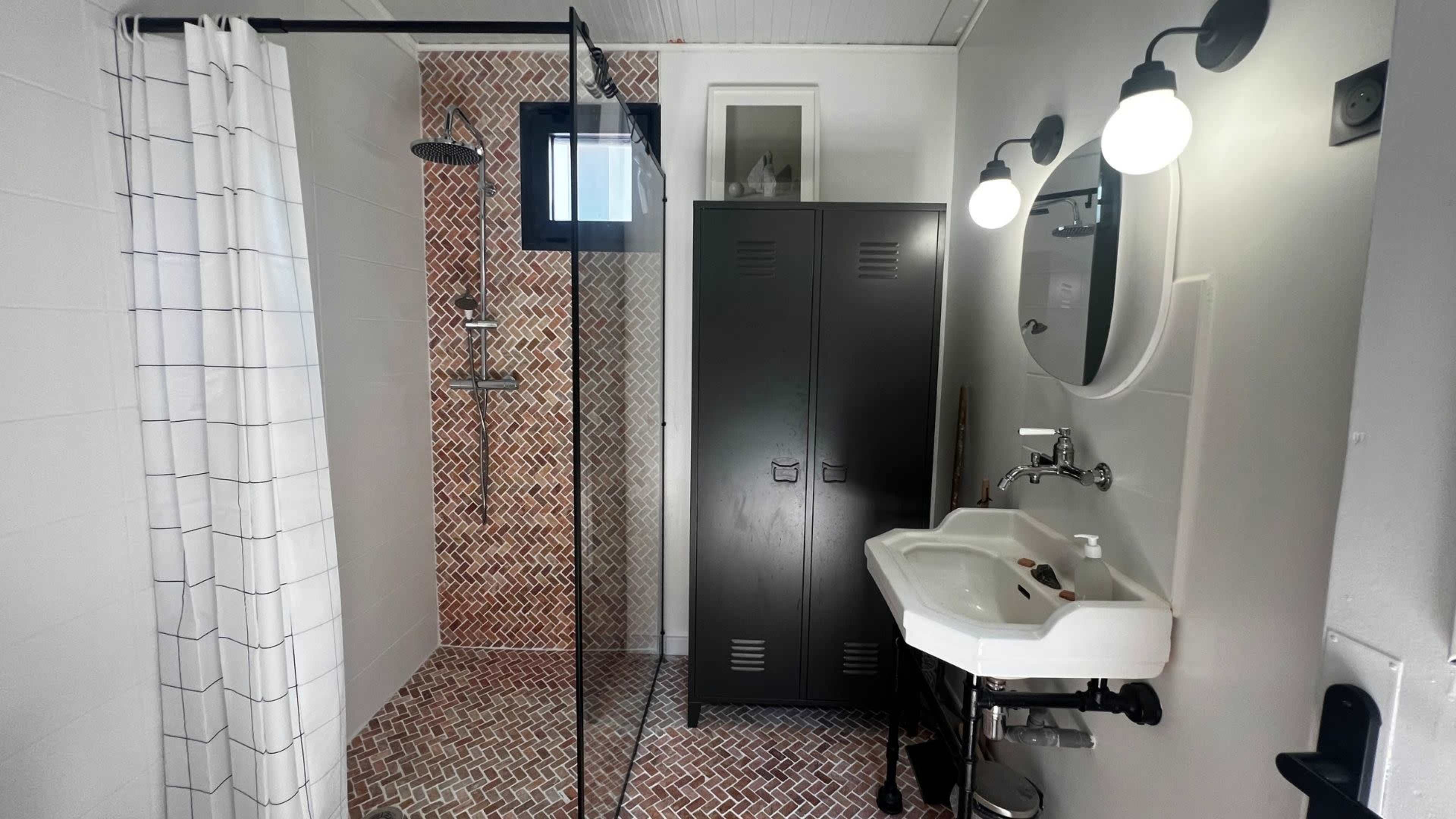The bathroom features a glass shower enclosure with a tiled wall, a large mirror with two sconces, and a sink next to a dark storage cabinet.