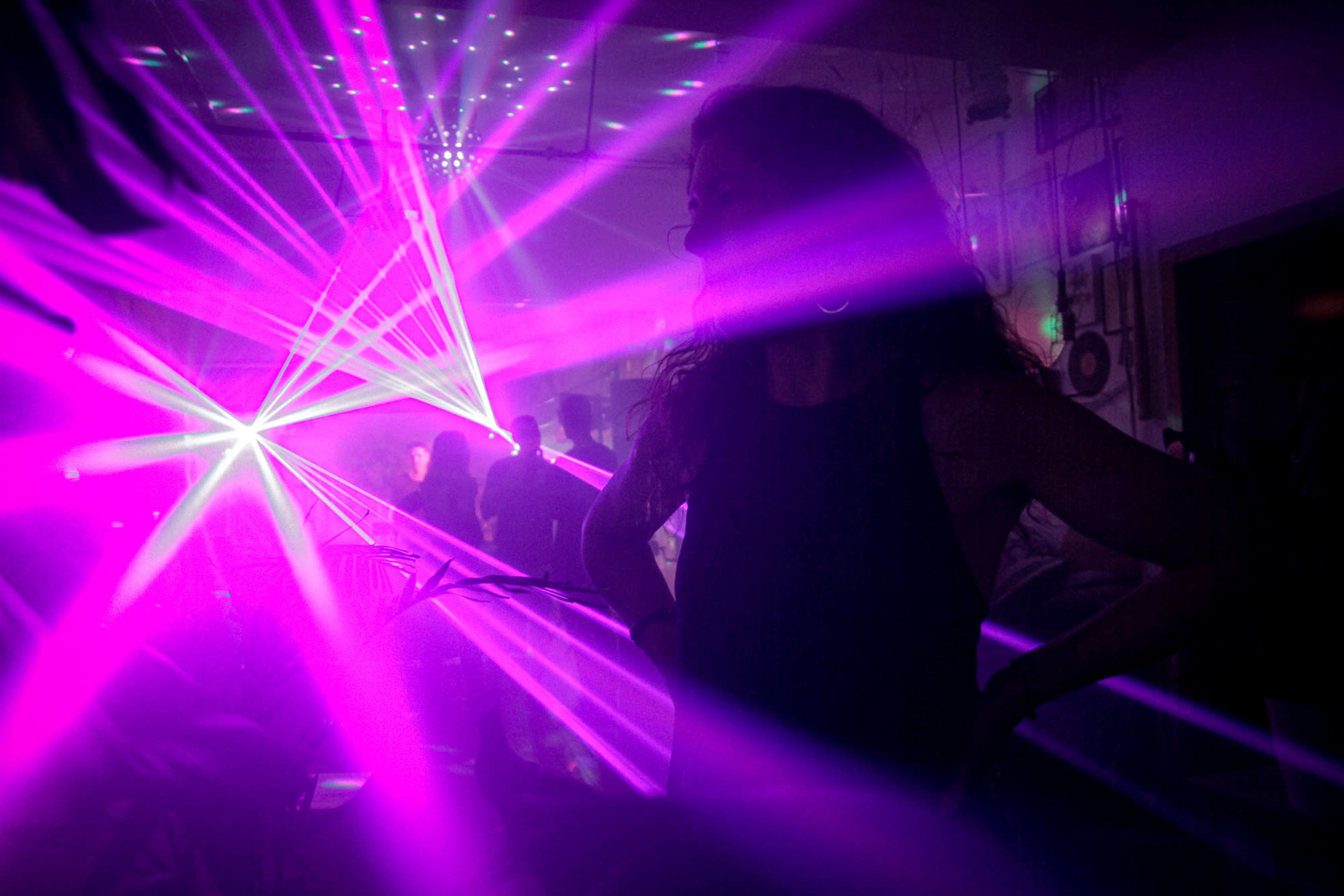 A woman stands with her hands on her hips in a dimly lit room filled with purple laser lights and silhouettes of people dancing.