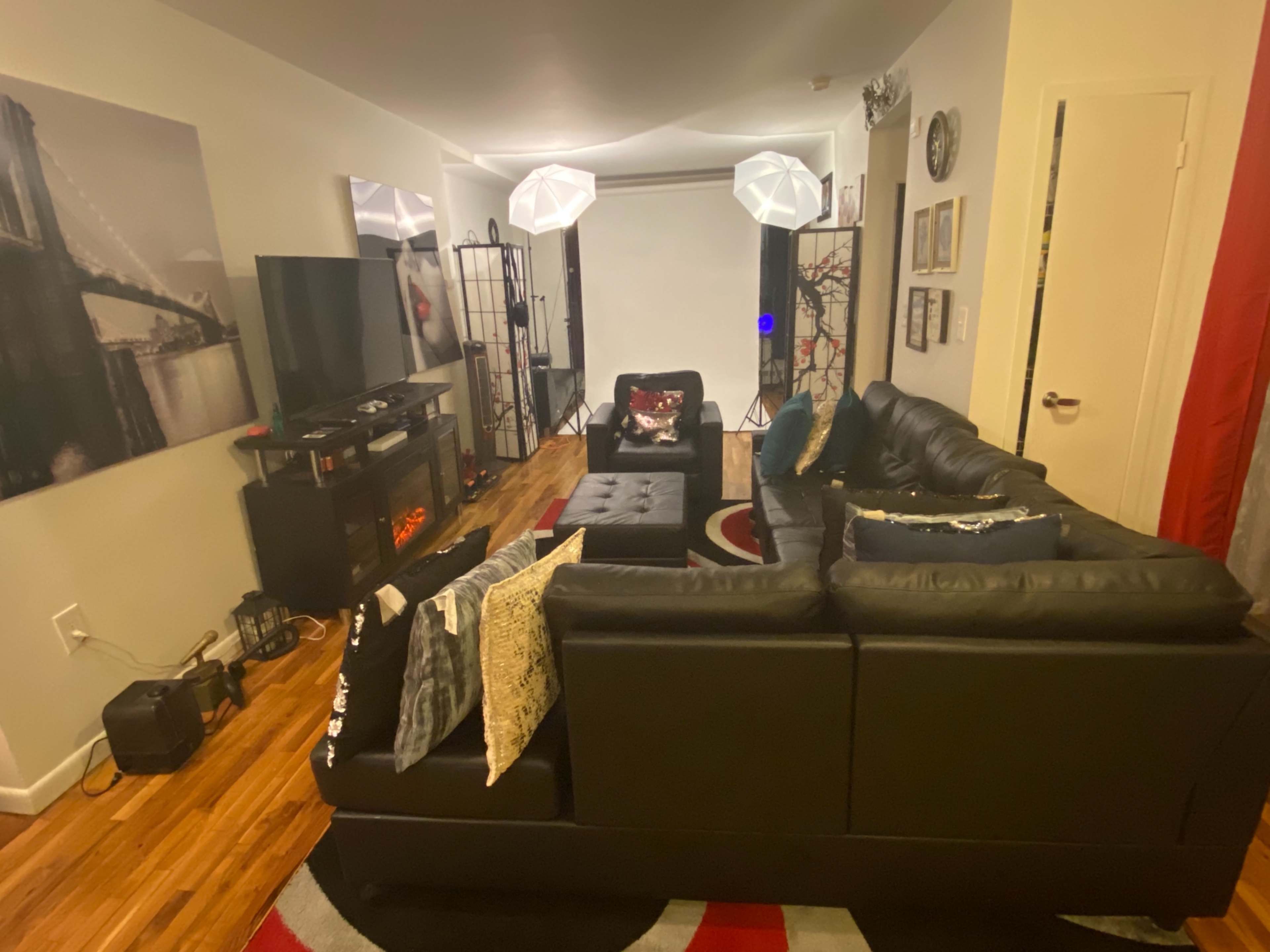 The image shows a living room with a black sectional sofa, a television, and decorative lighting, featuring a wooden floor and wall art.