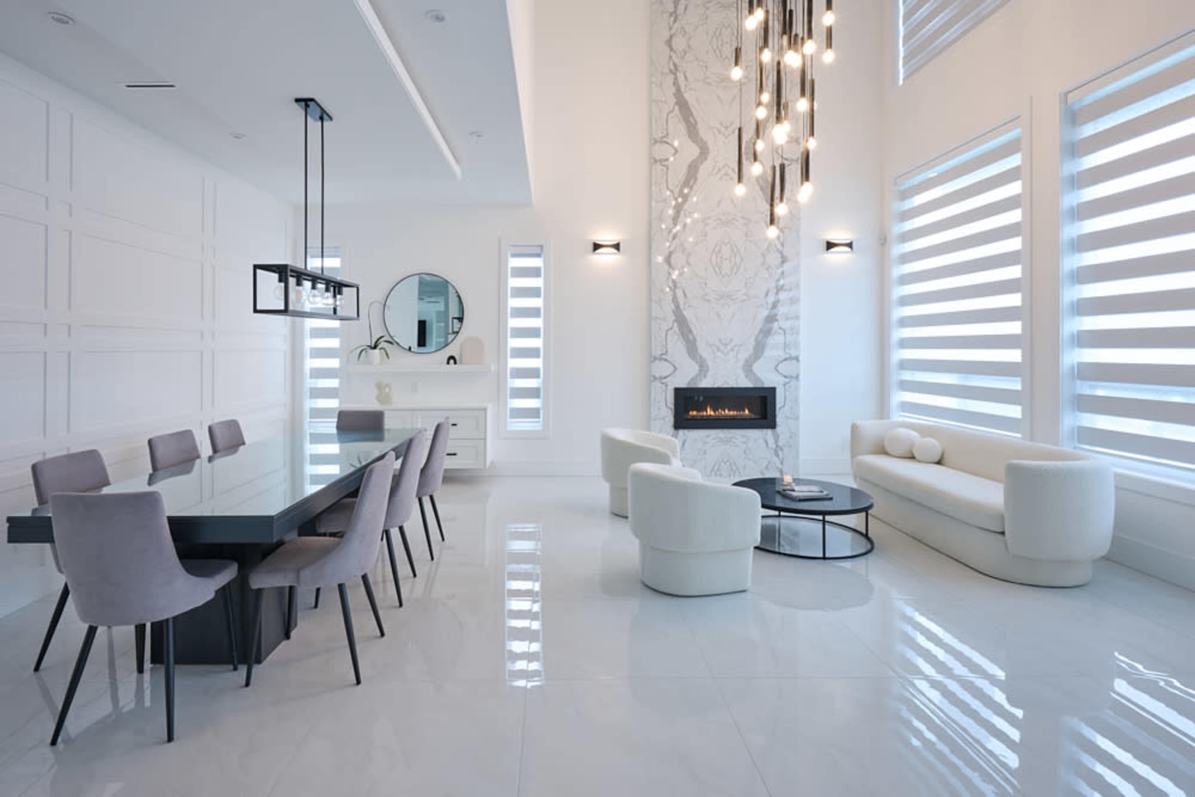 A modern living space featuring a dining area with a long table and upholstered chairs, a sleek fireplace along a patterned wall, and a minimalist seating area with a round coffee table.