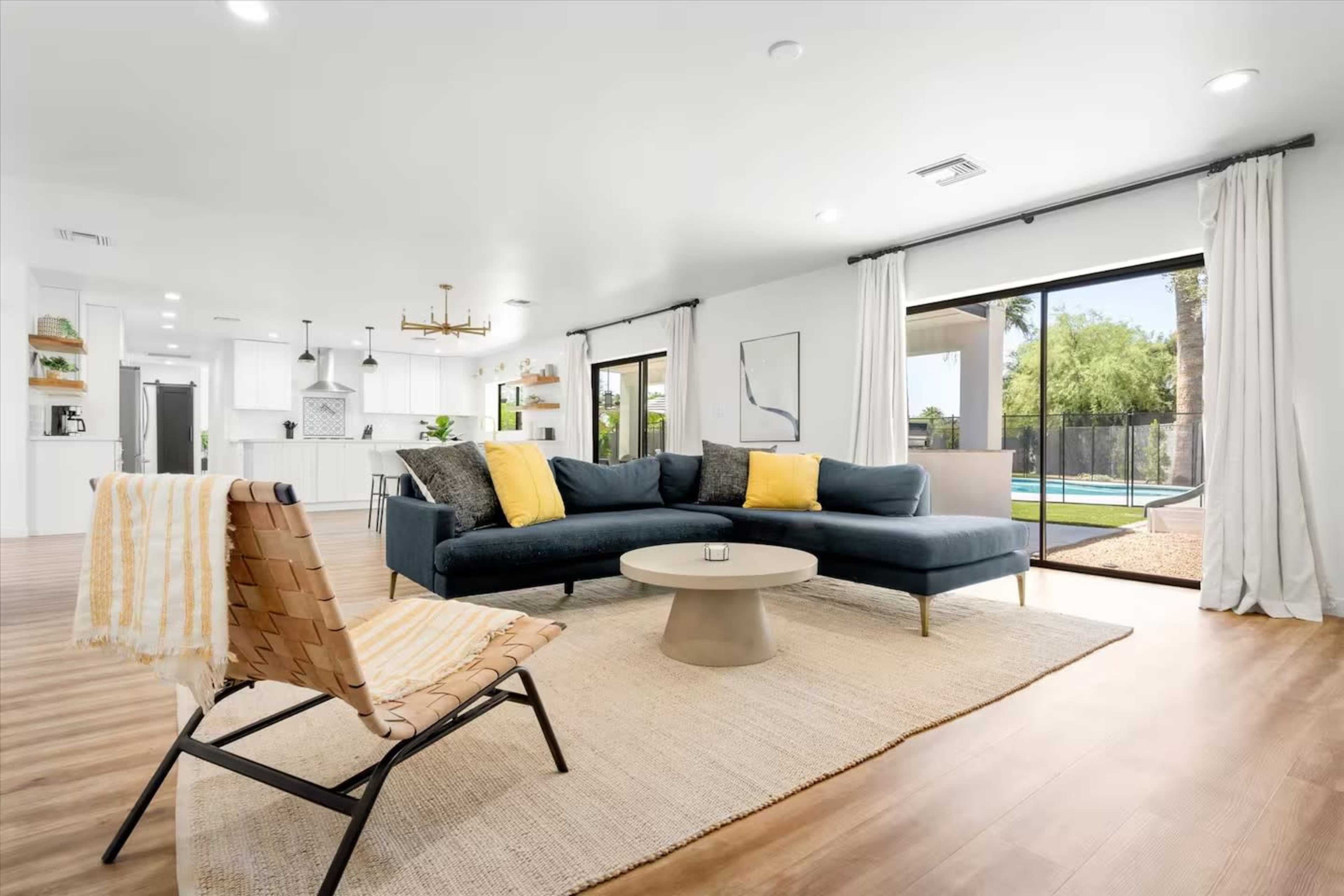 A spacious living room features a dark blue sectional sofa with yellow pillows, a round coffee table, and large sliding glass doors leading to a pool area.