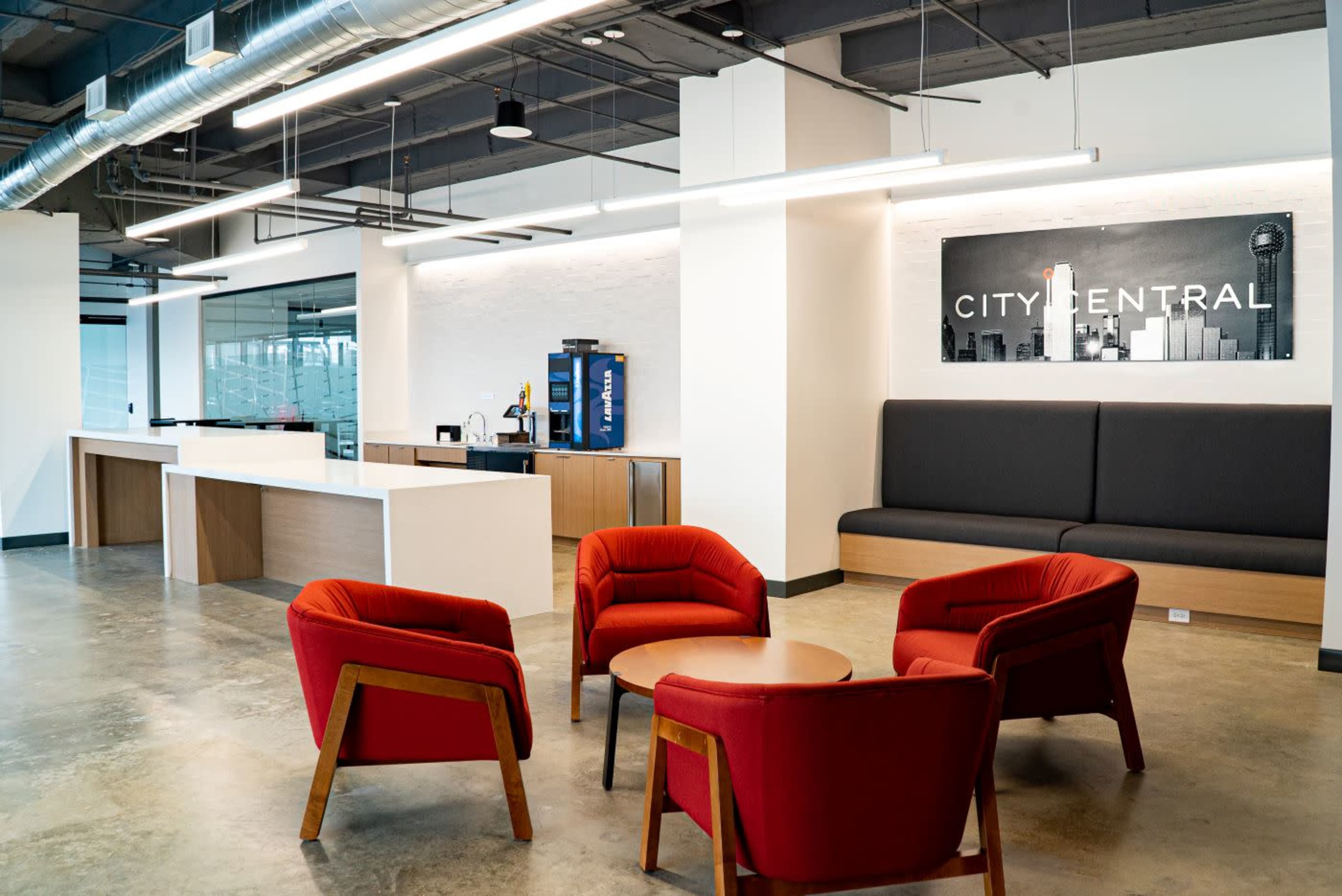 The image shows a modern office lobby with red armchairs surrounding a circular table, a wall display labeled "CITY CENTRAL," and a counter with a coffee station in the background.