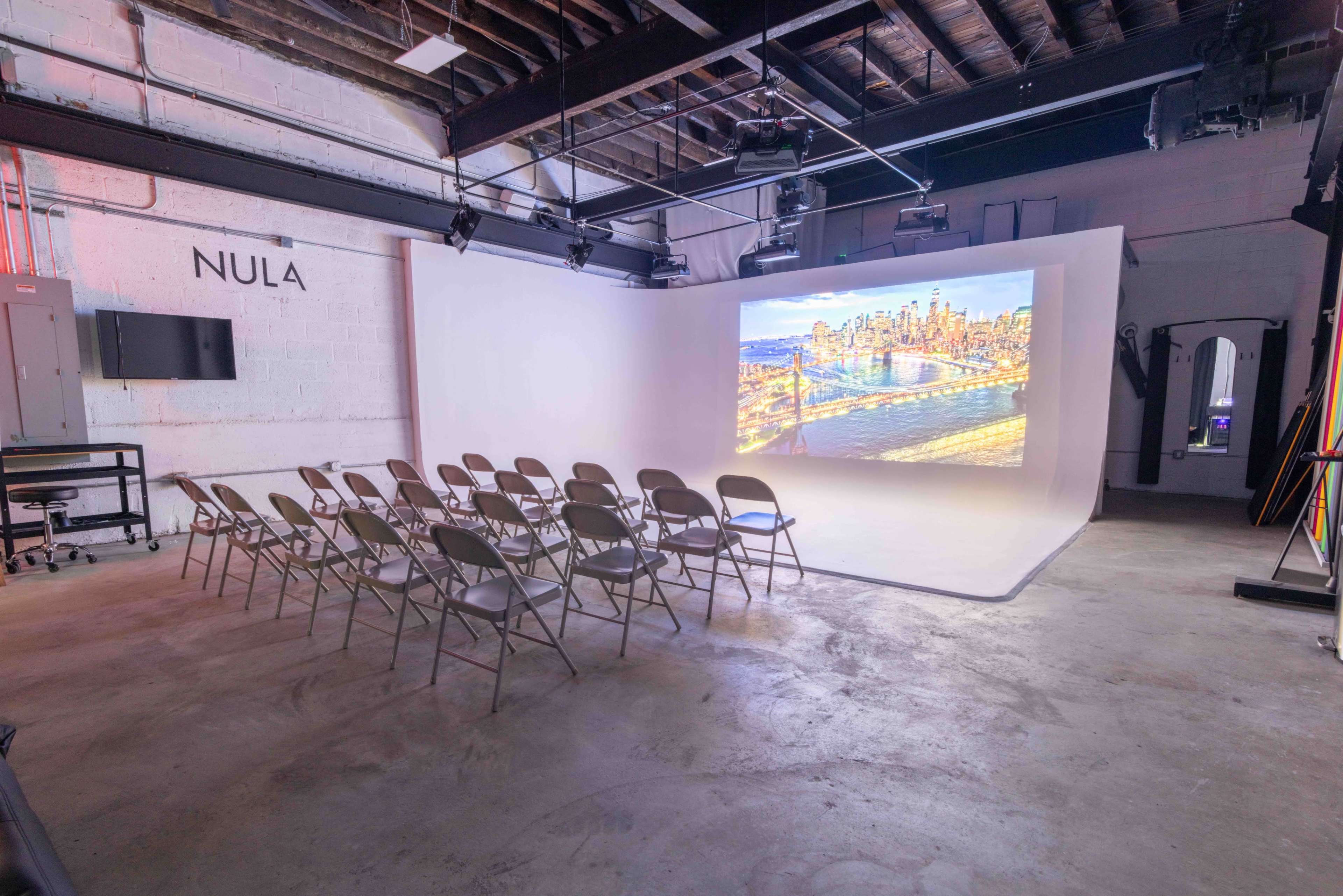 The image shows an empty interior space set up for presentations, featuring rows of folding chairs facing a large screen displaying a cityscape.