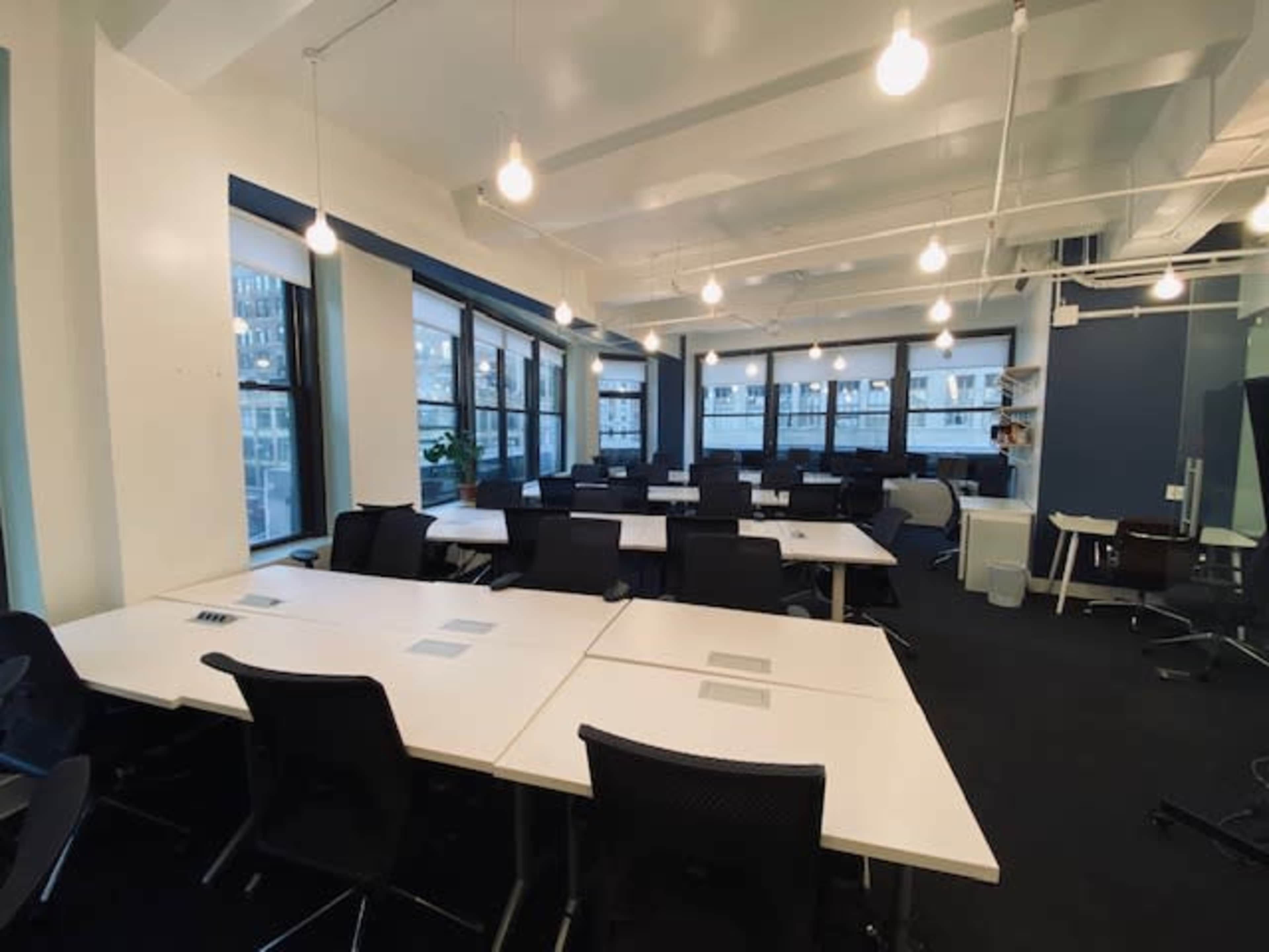 A modern office space with multiple empty desks arranged in rows, ample natural light coming through large windows, and overhead lighting.
