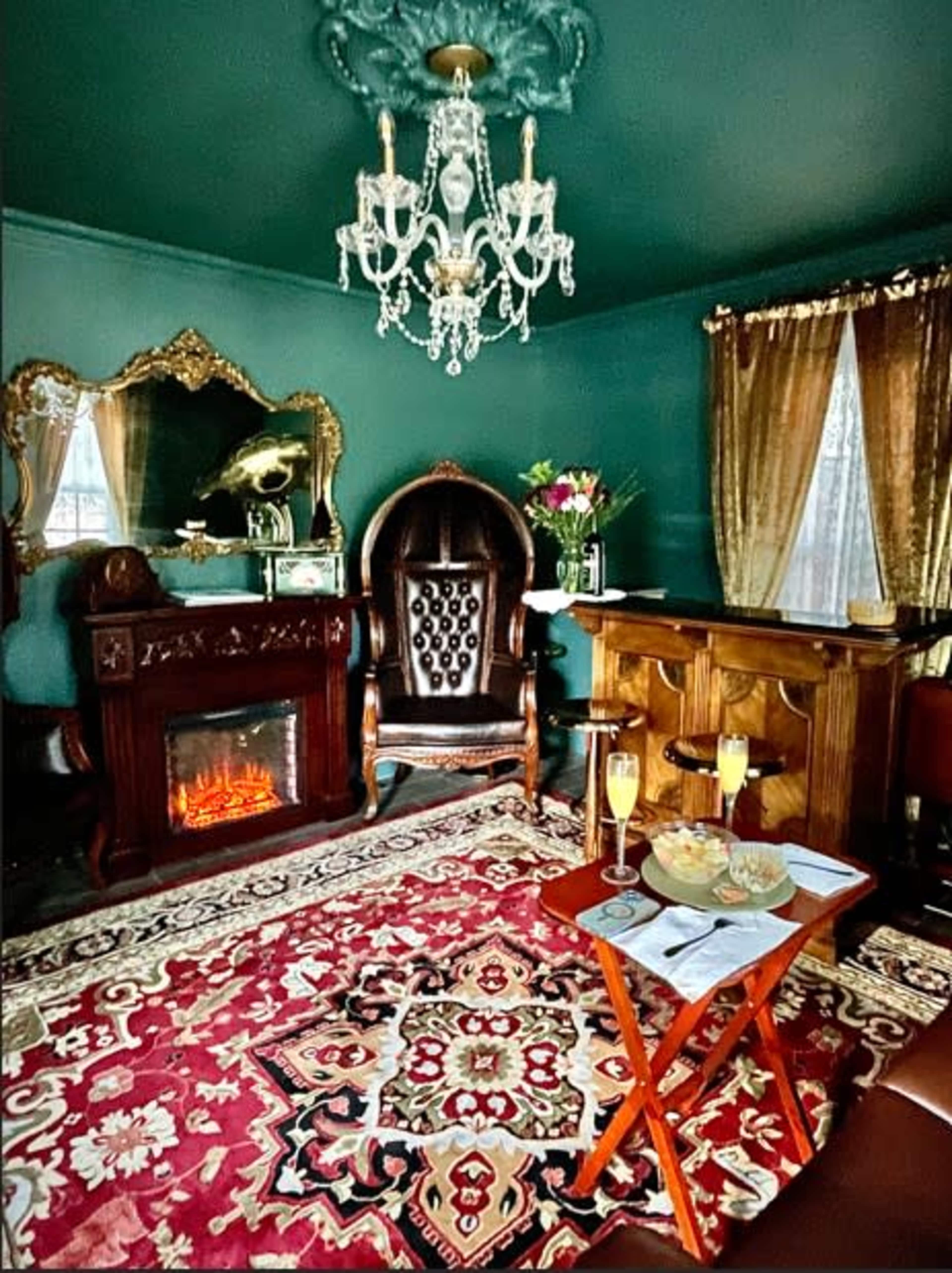 The image depicts a cozy room with a dark green wall, a fireplace, an ornate chandelier, and plush furnishings, including a large armchair and a small table with drinks and snacks.