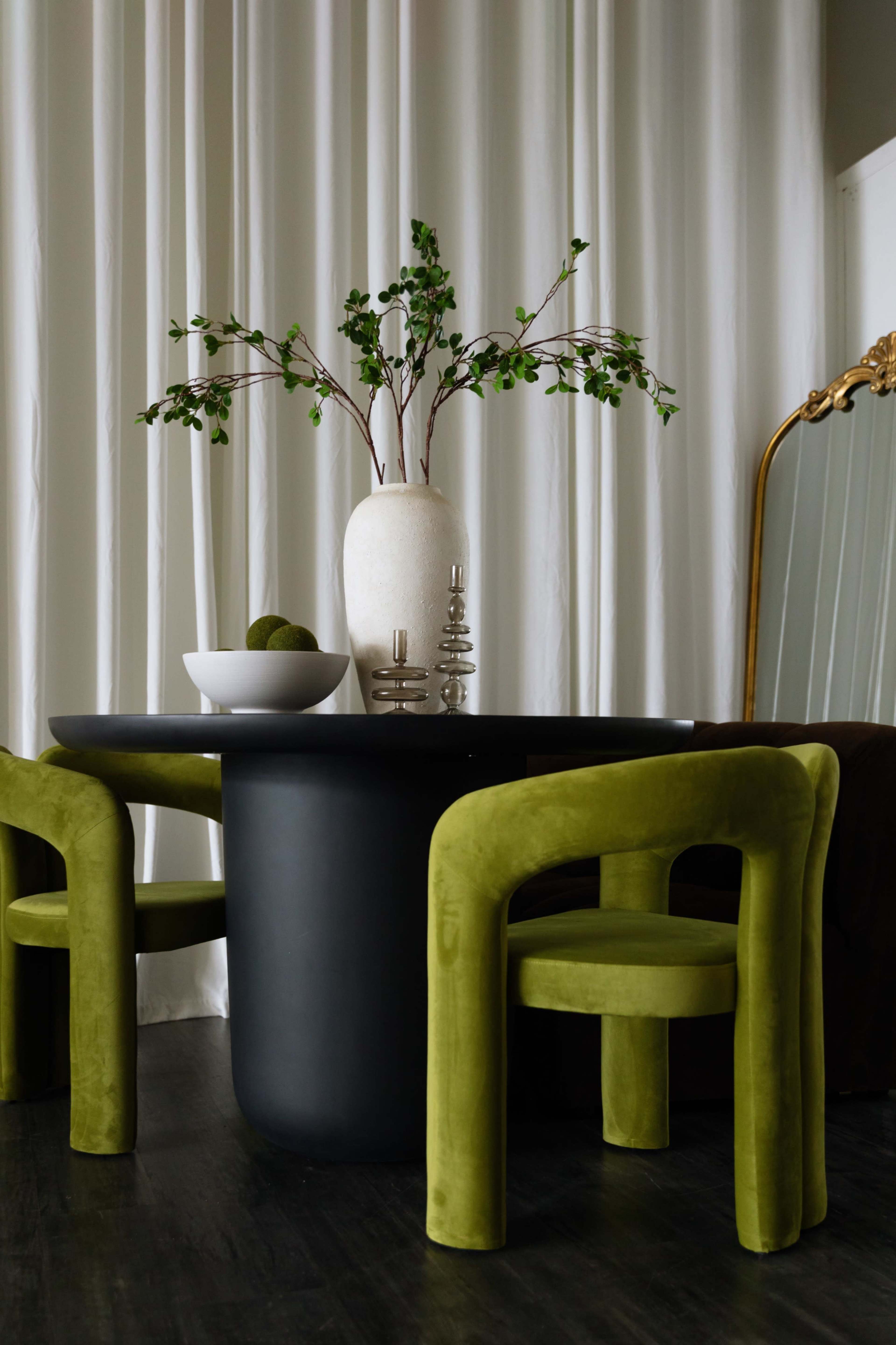 A round black table is set with a white vase containing branches and a bowl of green items, surrounded by two green upholstered chairs against a backdrop of white curtains.