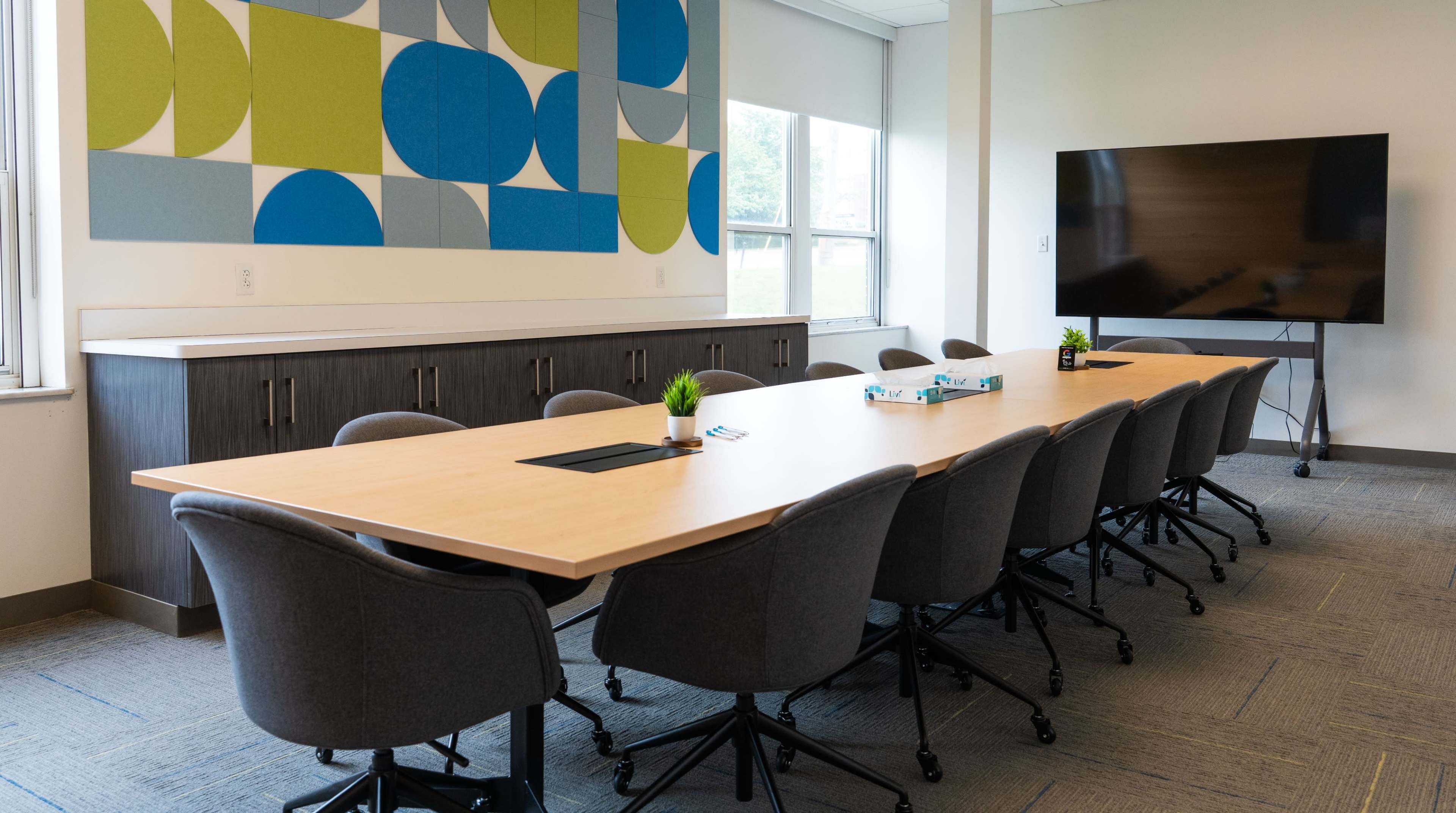A modern conference room features a long wooden table surrounded by rolling chairs, with a large screen and a colorful wall mural in the background.