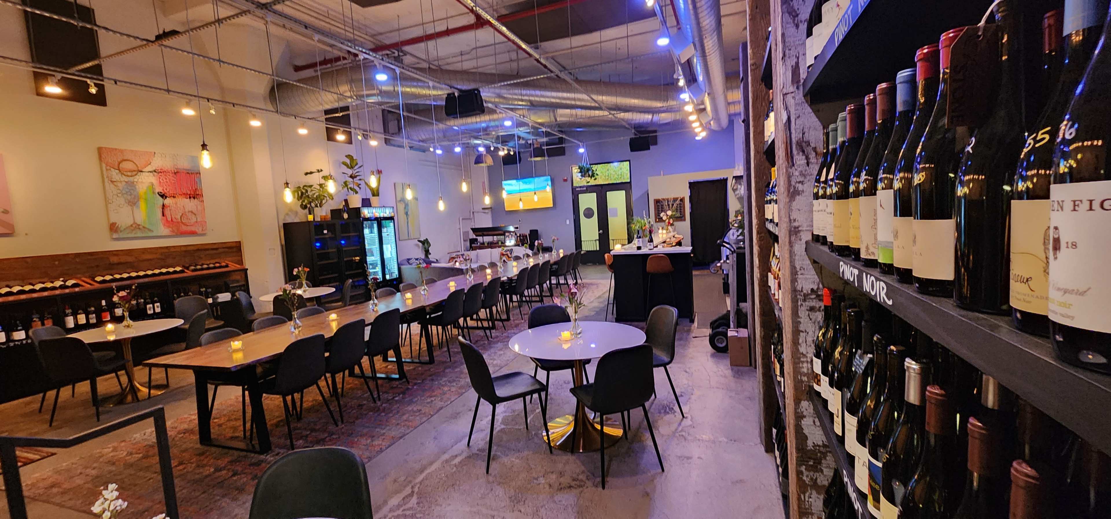 The image shows a modern wine bar with a long table and chairs in the center, flanked by shelves of wine bottles on one side and a bar area on the other.