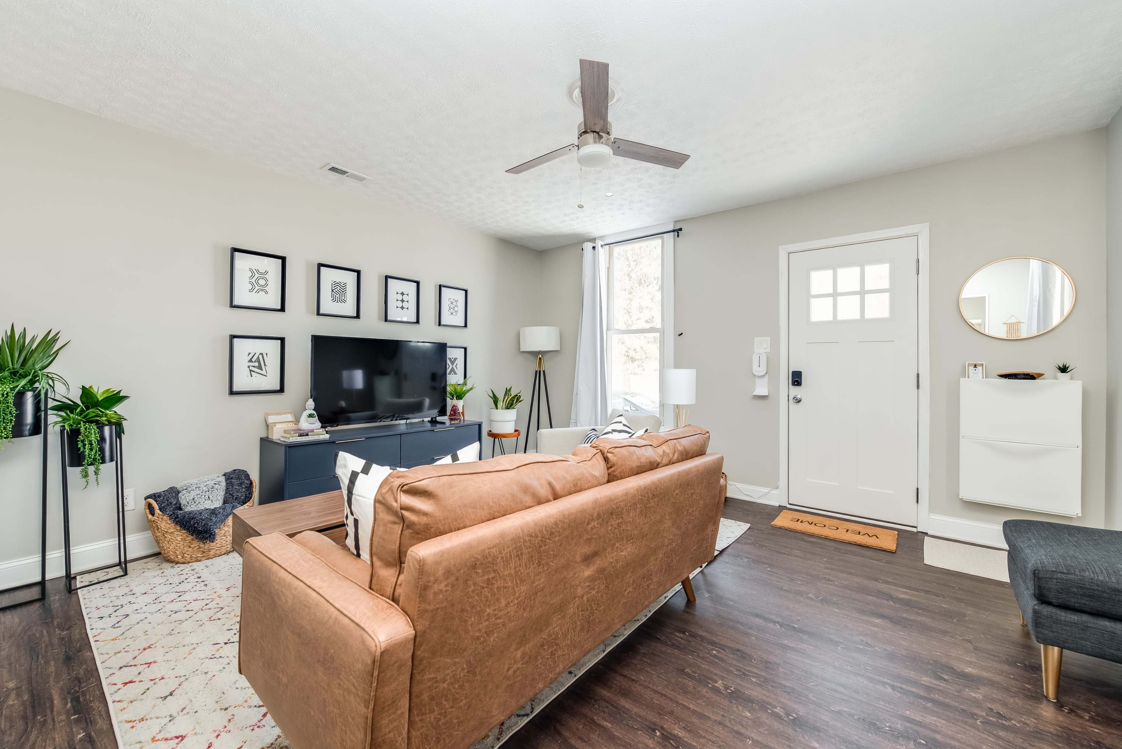 The living room features a brown leather couch, a television on a navy console, and large windows with light curtains, accompanied by several plants and modern decor.