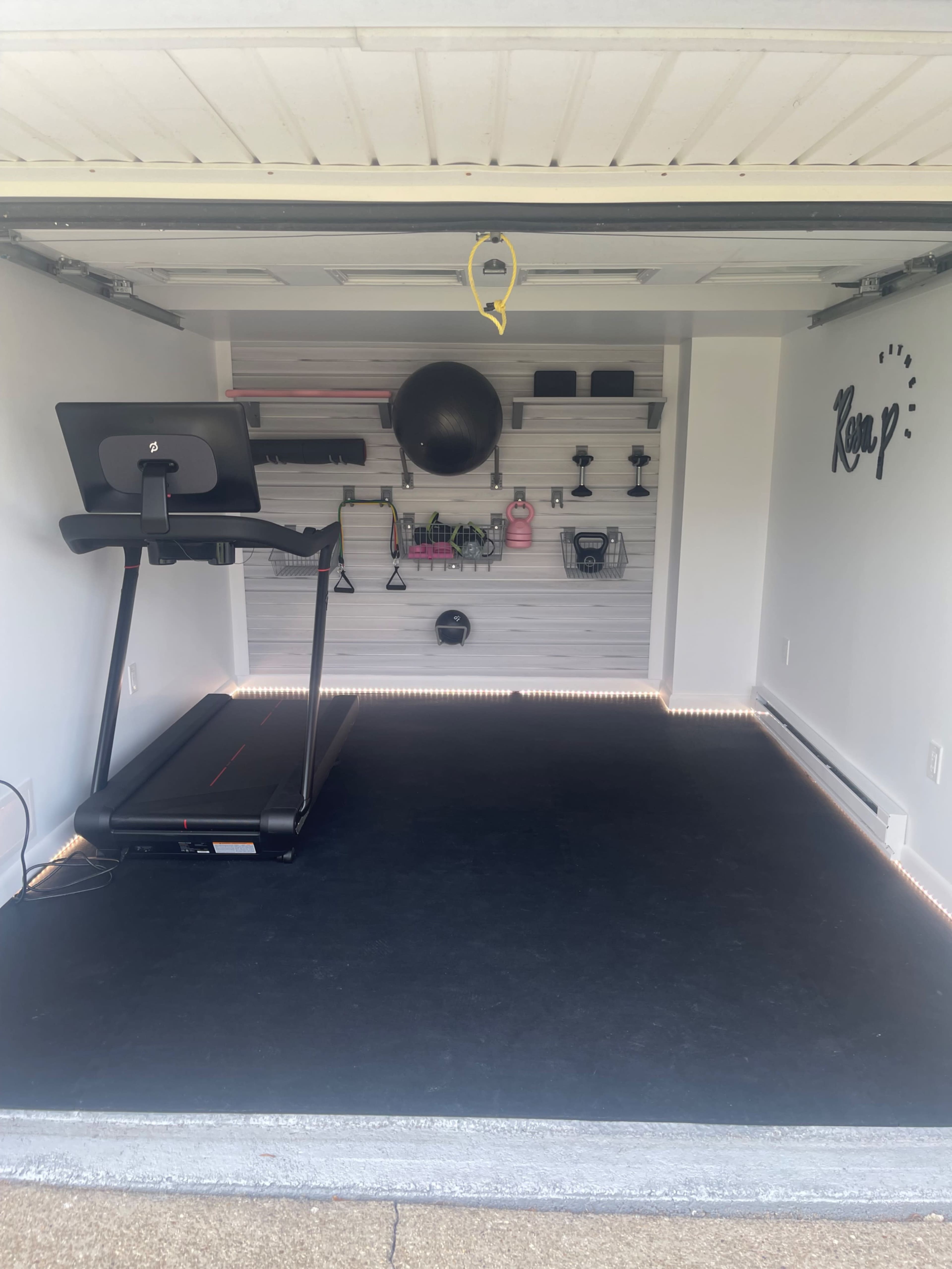 A home gym setup in a garage, featuring a treadmill, various exercise equipment on shelves, and a rubber flooring.