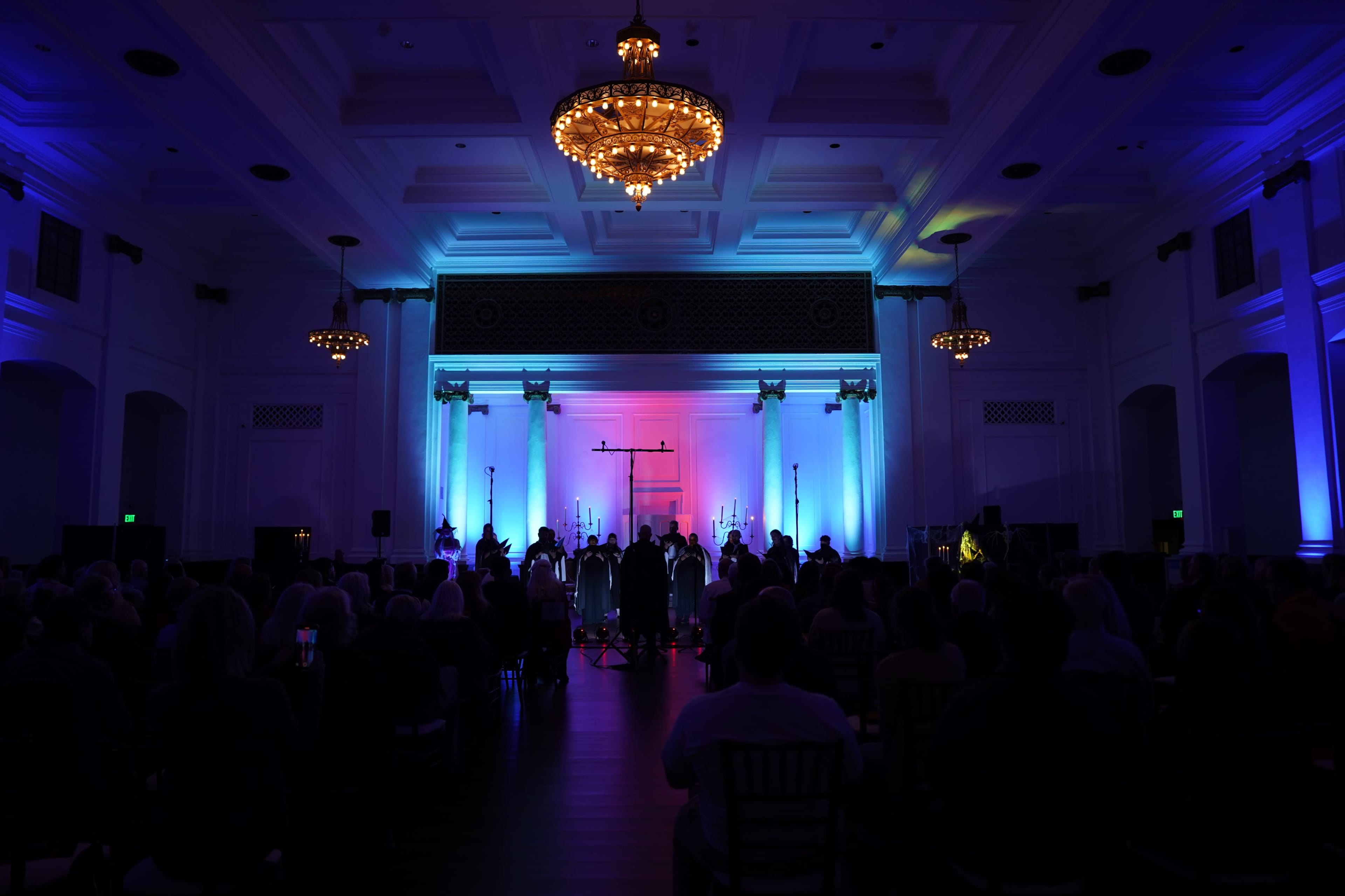 A stage is set in a large hall, illuminated with colorful lighting, featuring a band performing in front of an audience seated at tables.