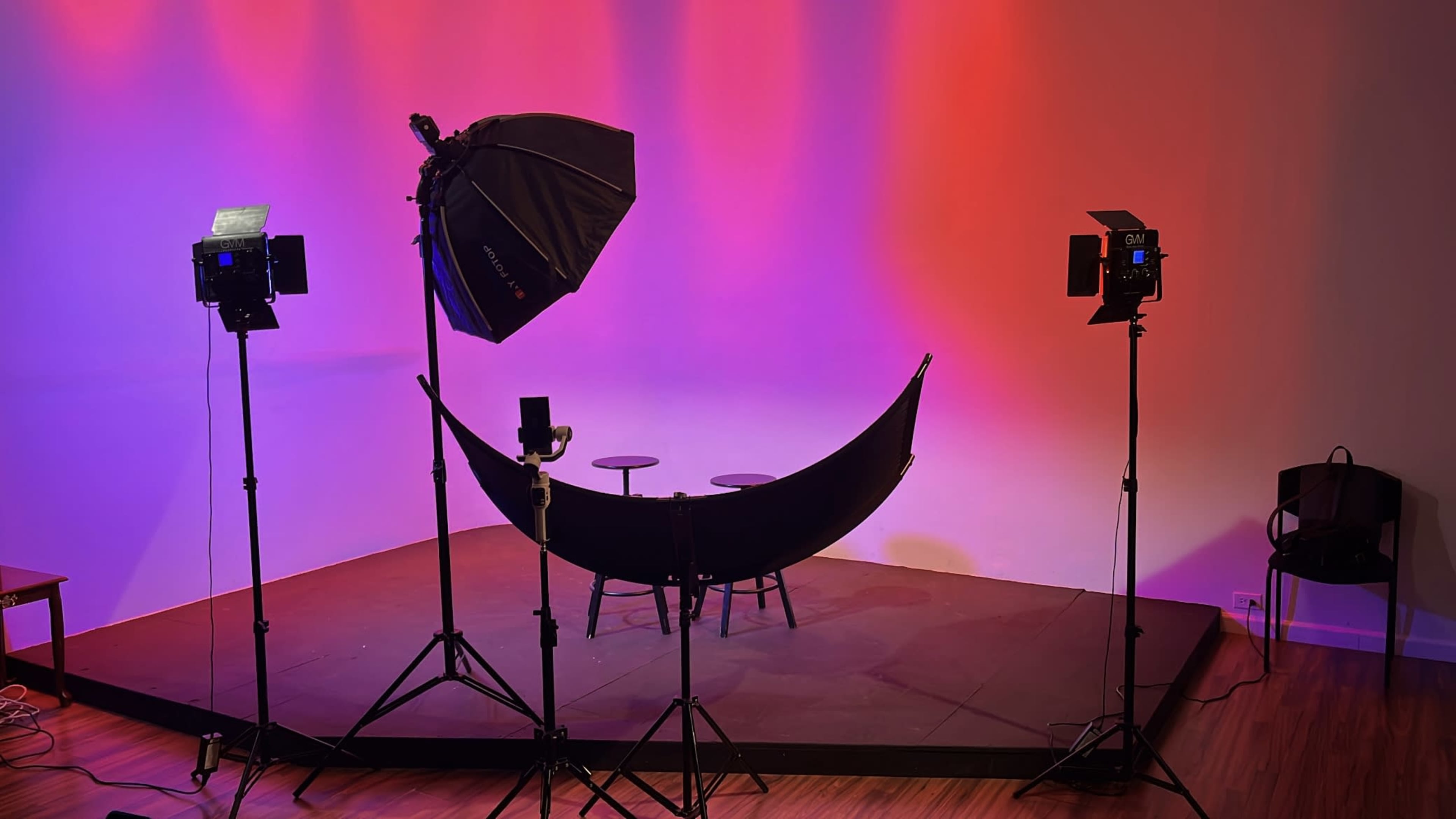 The image shows a photography studio setup with two light stands, a large softbox, a backdrop, and two stools on a raised platform.