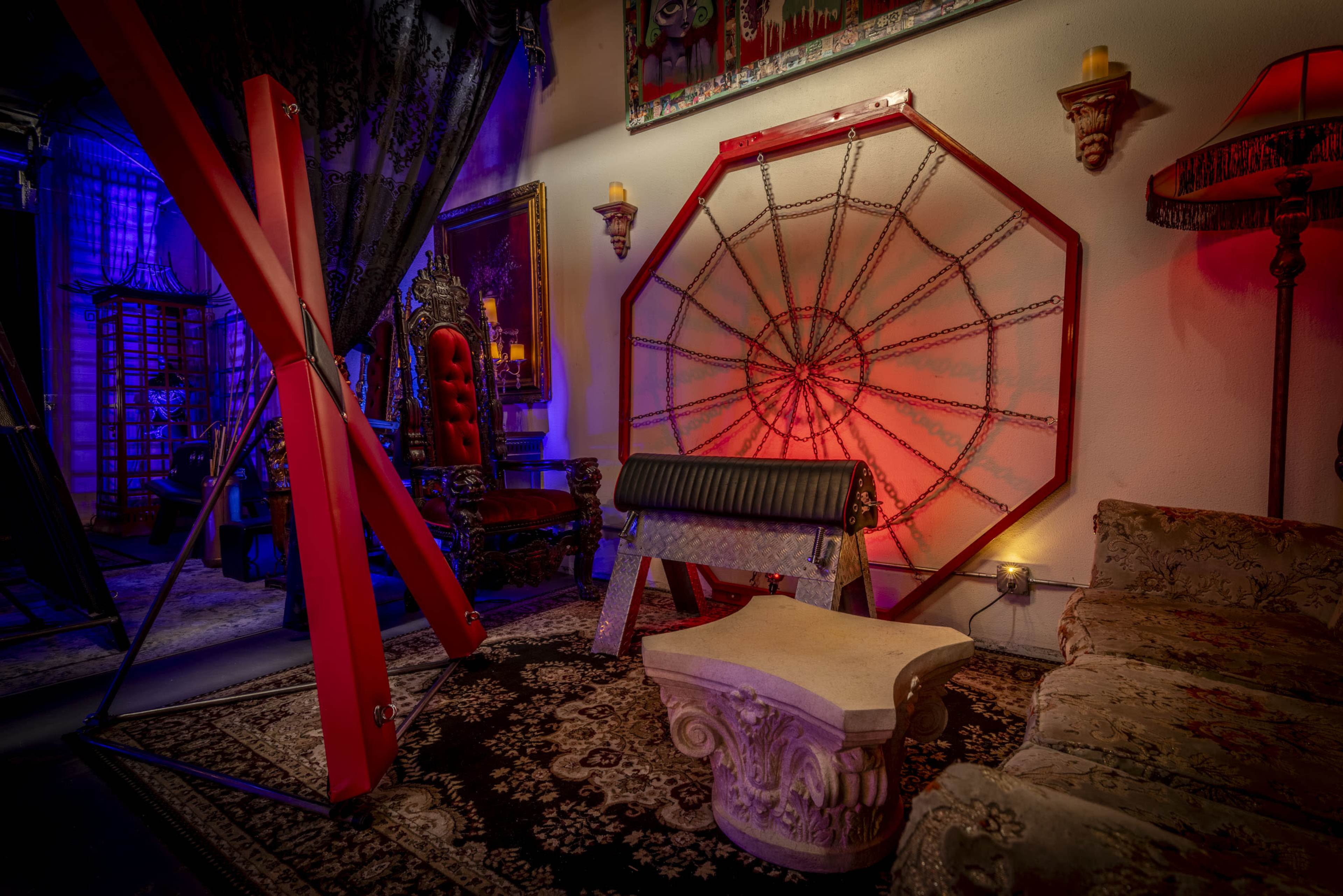 The scene features a dimly lit room with ornate furniture, including a couch and a sculpted table, alongside a large, red spider web structure and BDSM equipment.