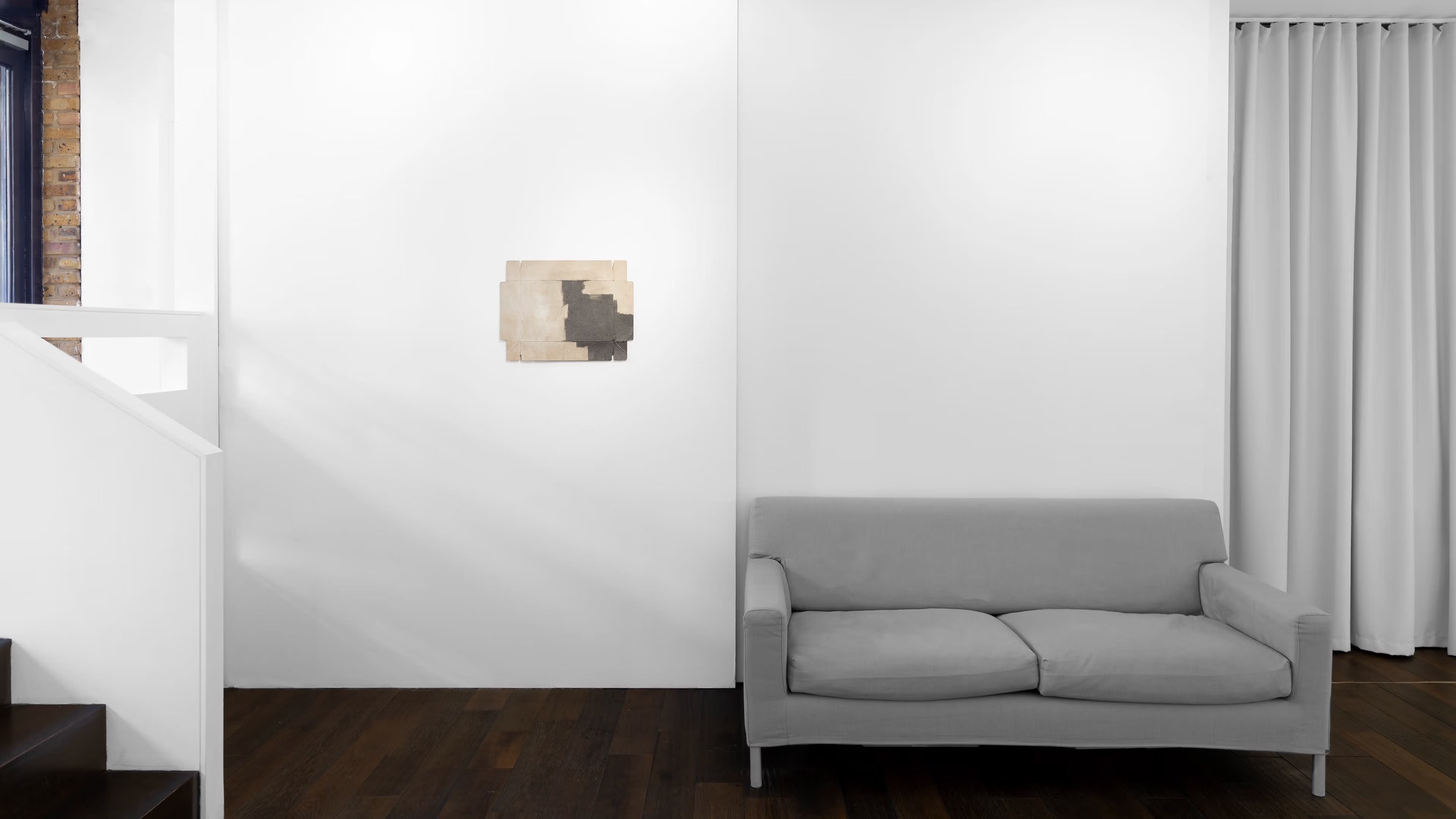 A gray sofa is positioned against a white wall, which features a small abstract artwork, alongside a pair of white curtains.