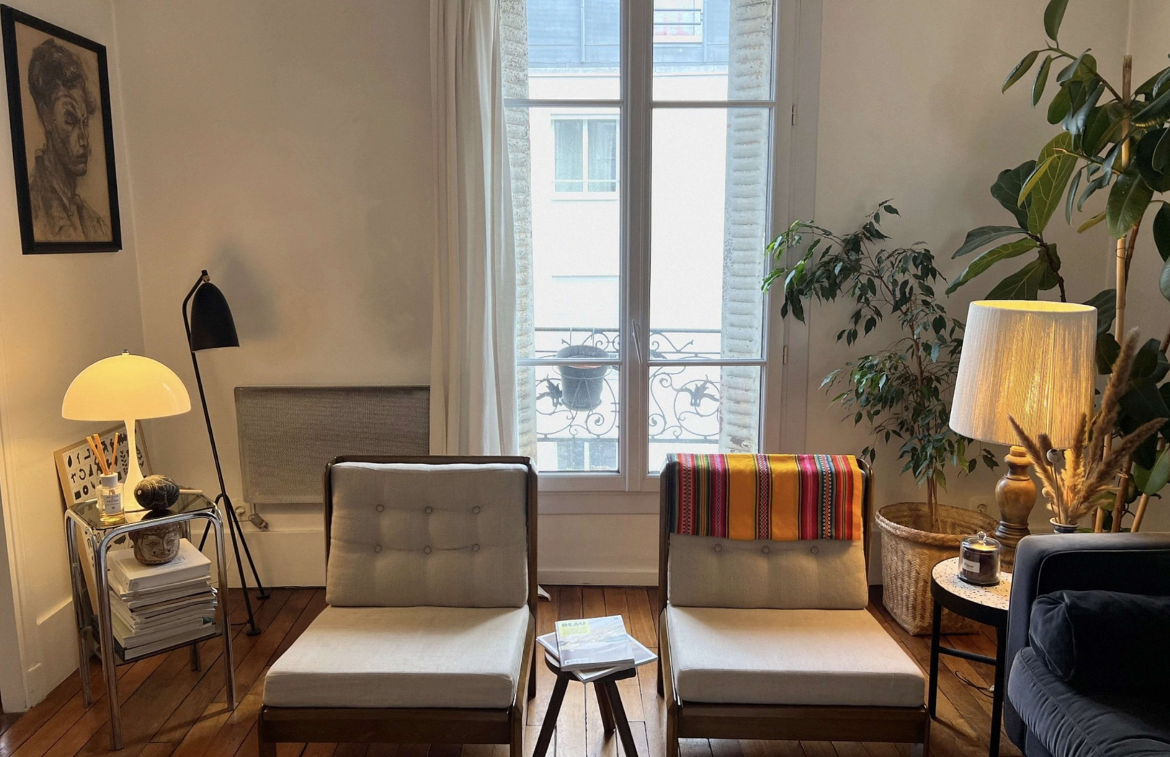 The image shows a cozy living room with two armchairs facing a window, a floor lamp, and decorative plants.