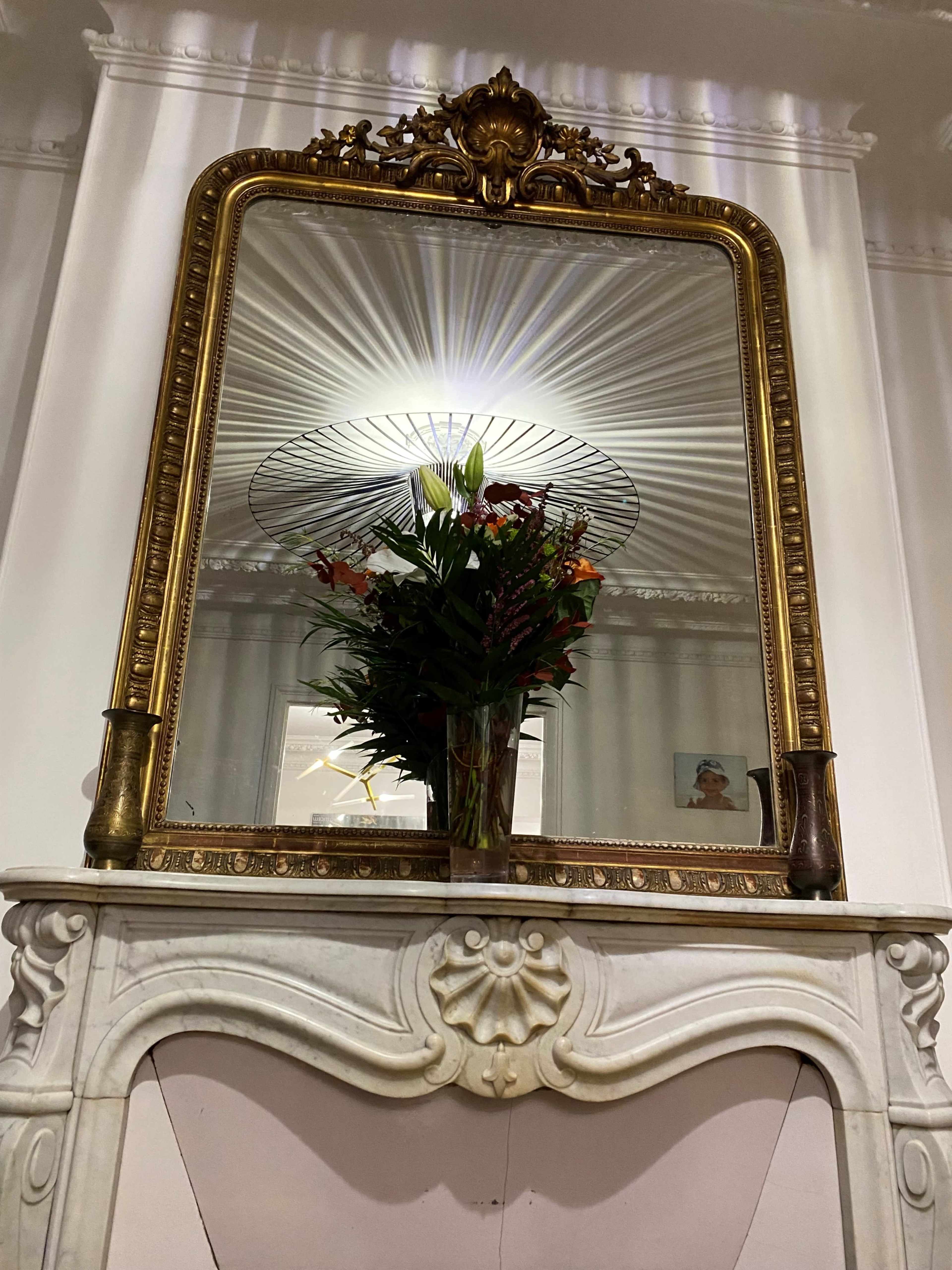 A large ornate mirror is positioned above a marble fireplace, reflecting a bouquet of flowers and creating a starburst effect from overhead lighting.