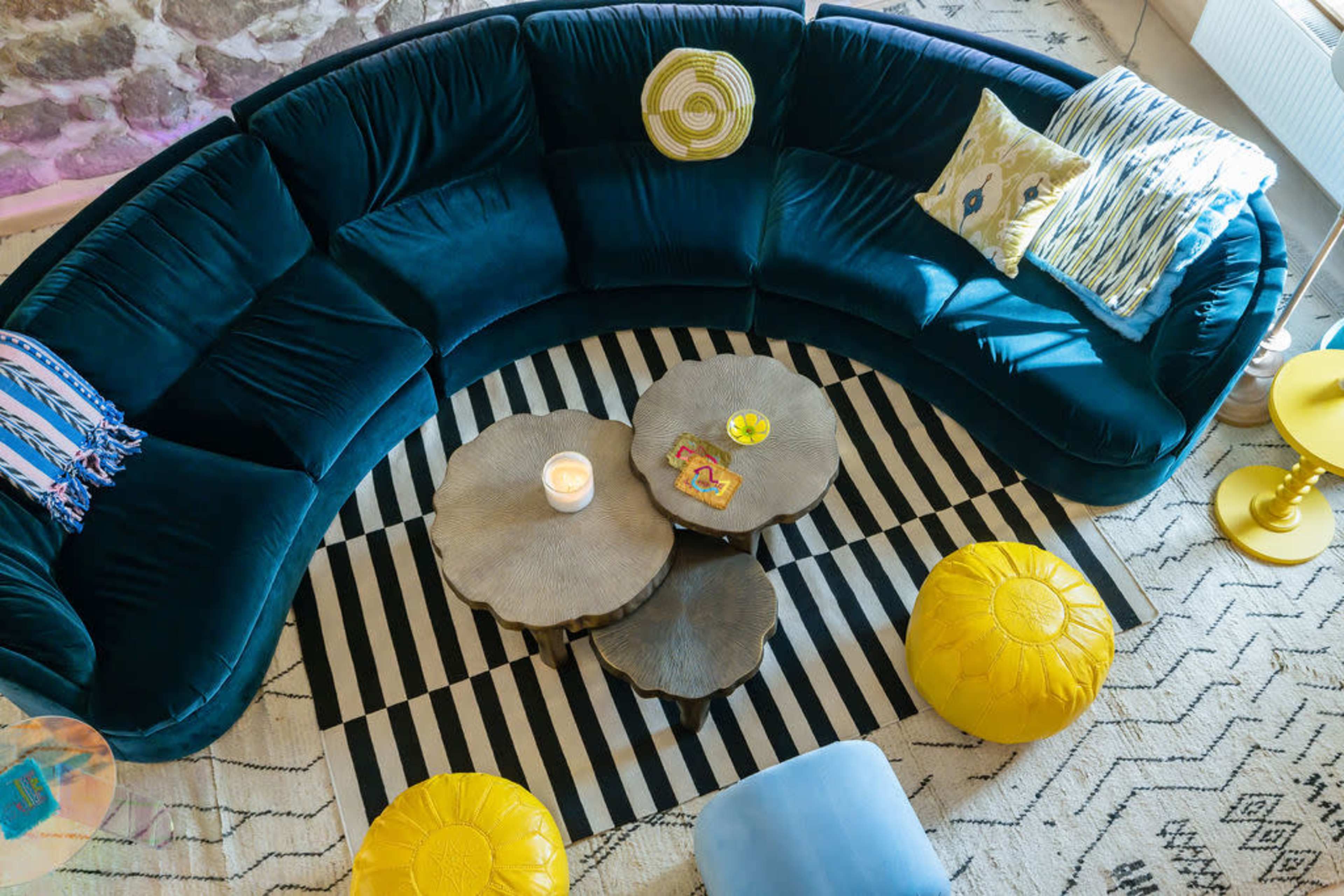 A circular, dark blue velvet sofa arranged around two tree stump-style coffee tables on a striped rug, with yellow poufs and decorative pillows scattered around.