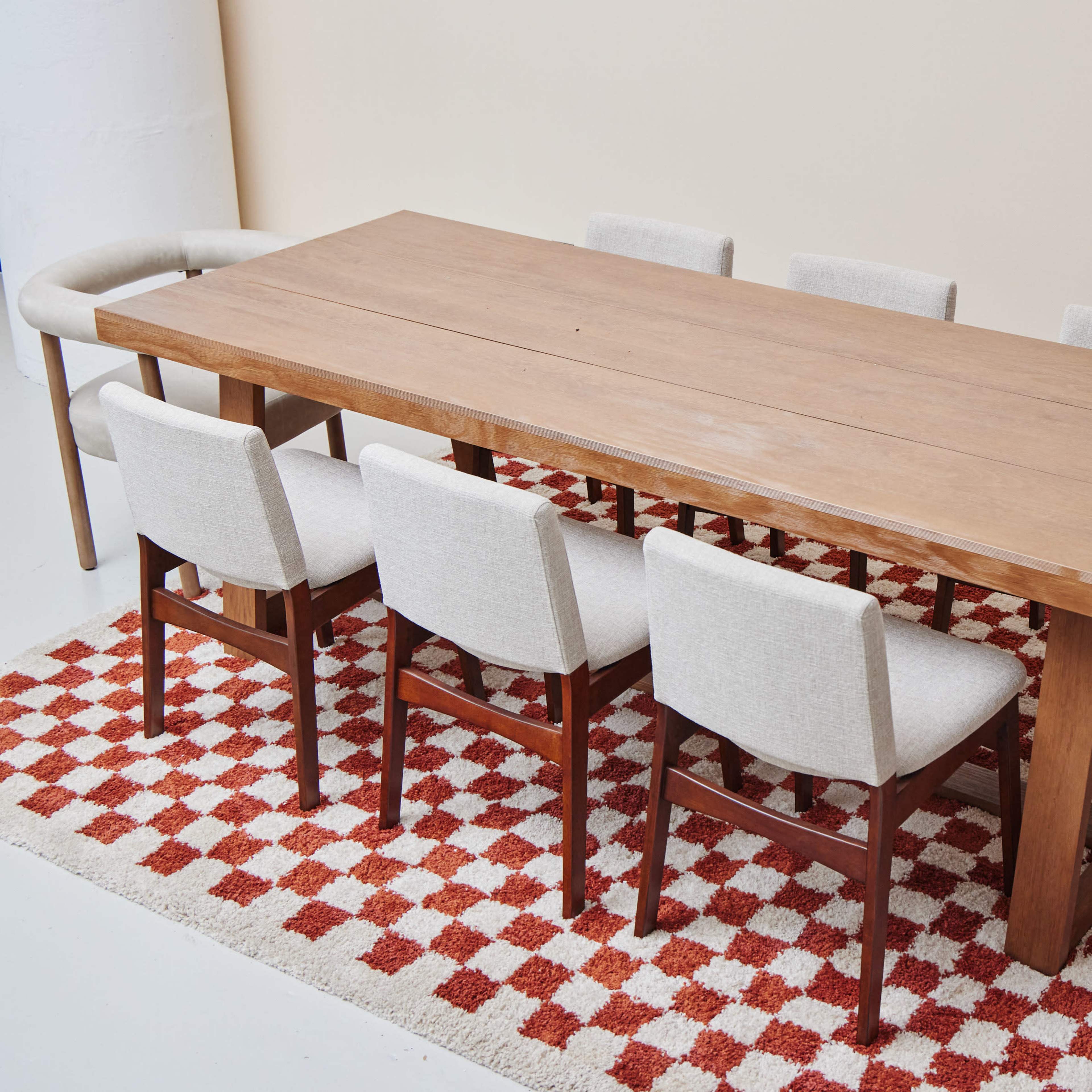 A wooden dining table with a smooth surface is surrounded by six upholstered chairs on a red and white checkered rug.