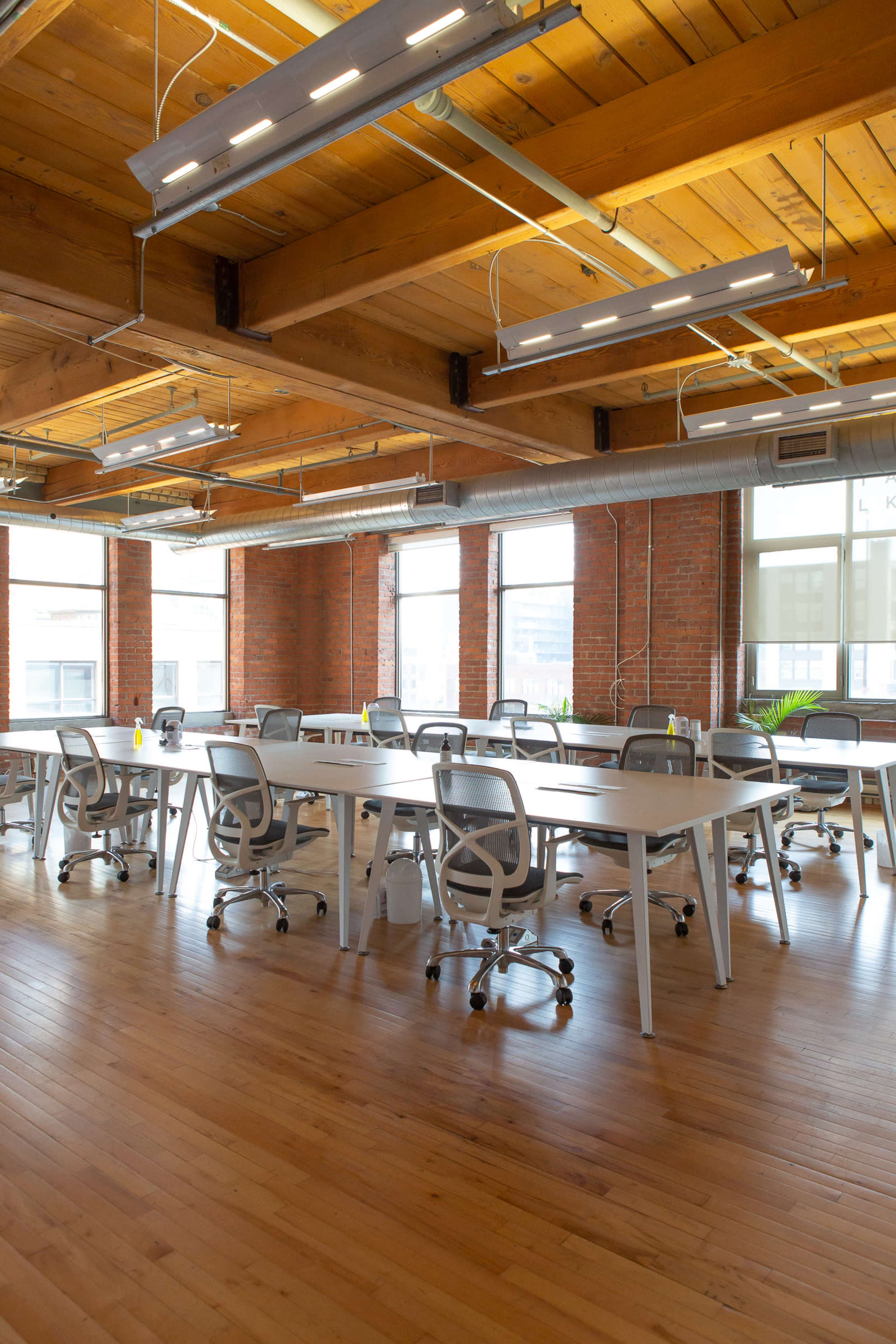 A spacious office with wooden floors and large windows, featuring multiple work tables and ergonomic chairs arranged in rows.
