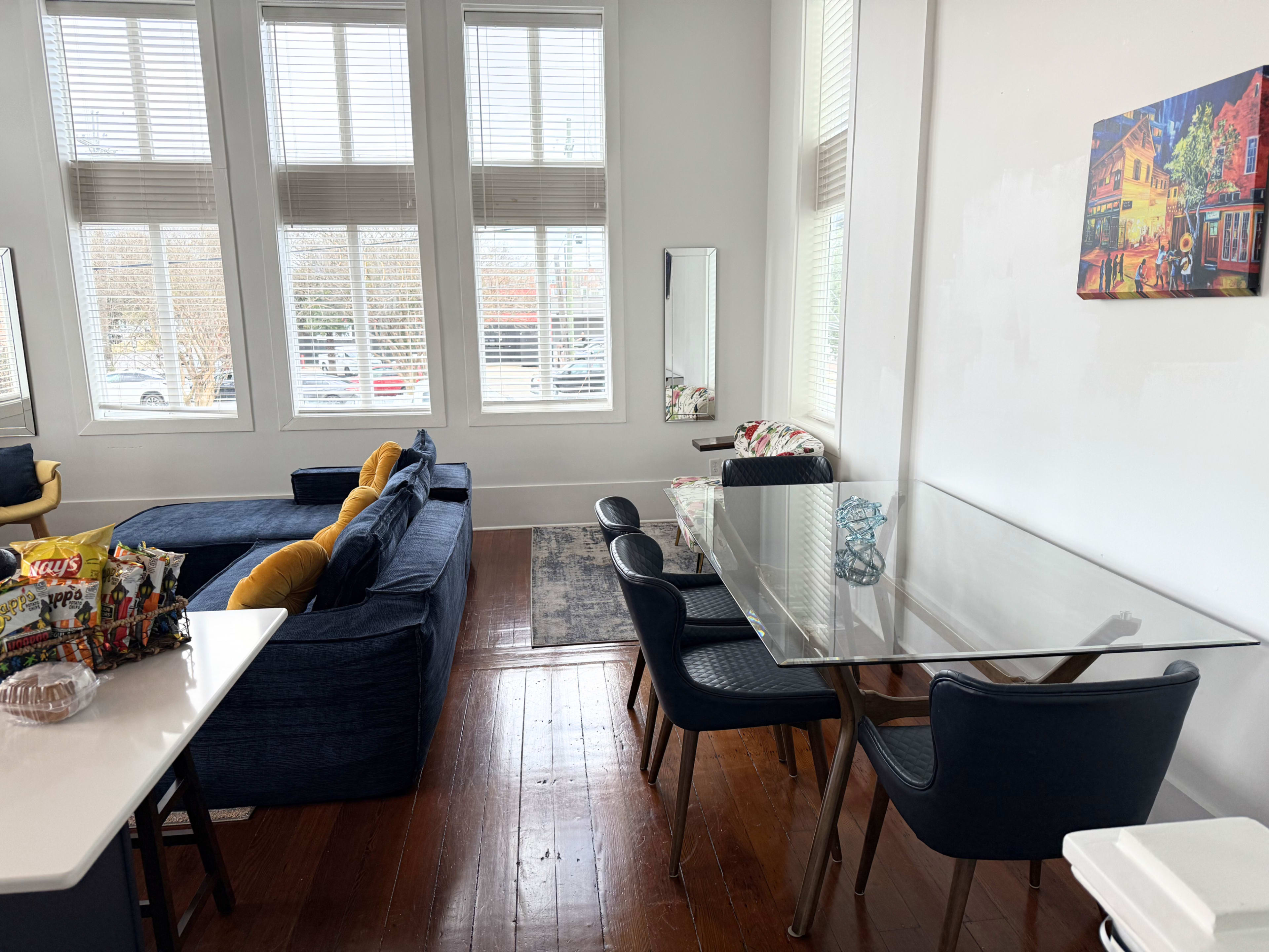 The image shows a modern living space featuring a blue sofa, a glass dining table with black chairs, and large windows allowing natural light to enter.