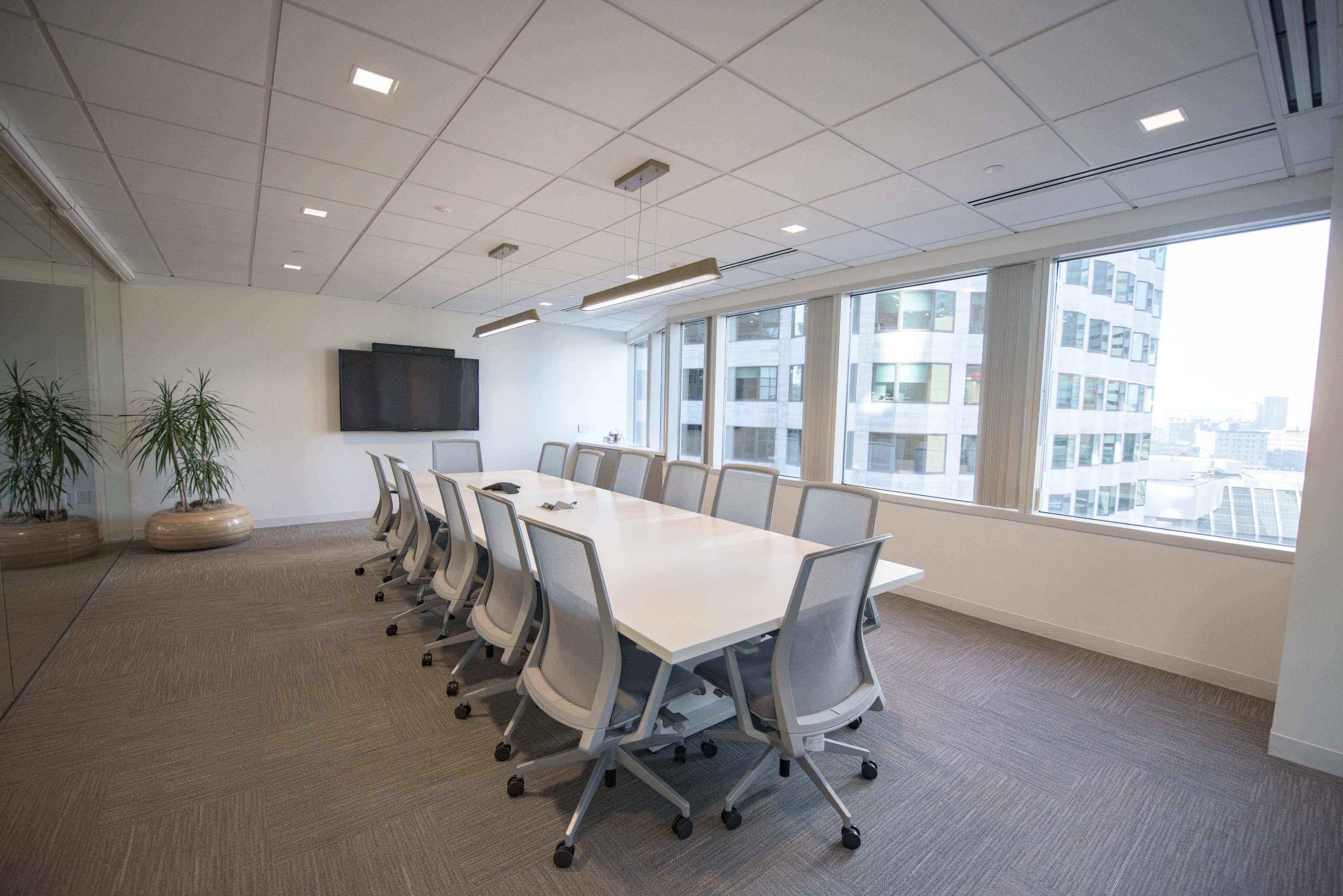 A conference room featuring a long white table, ergonomic chairs, large windows, and a wall-mounted screen.