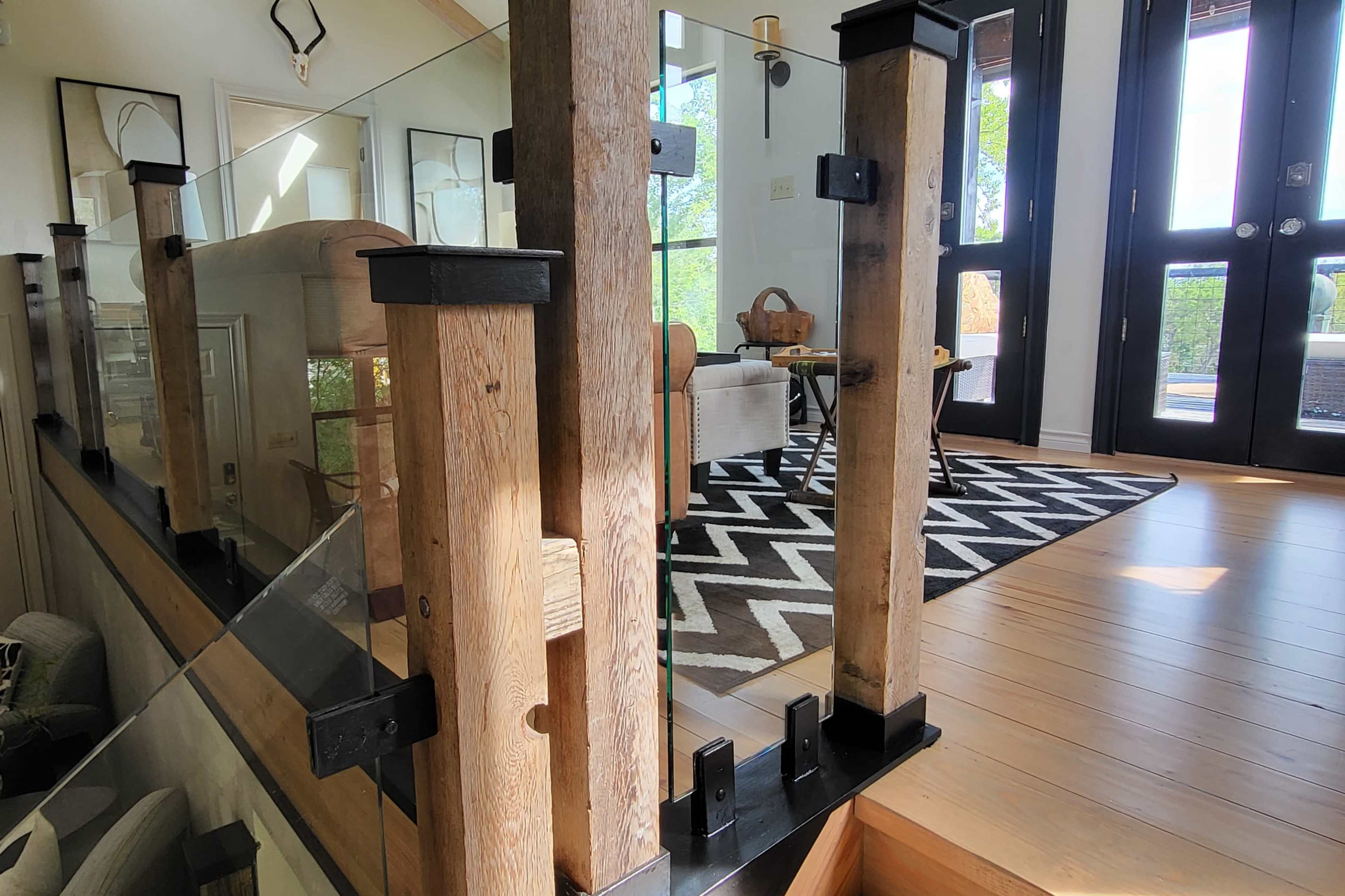 The image shows a modern interior space featuring wooden posts and glass railing, with a striped rug and furniture visible in the background.