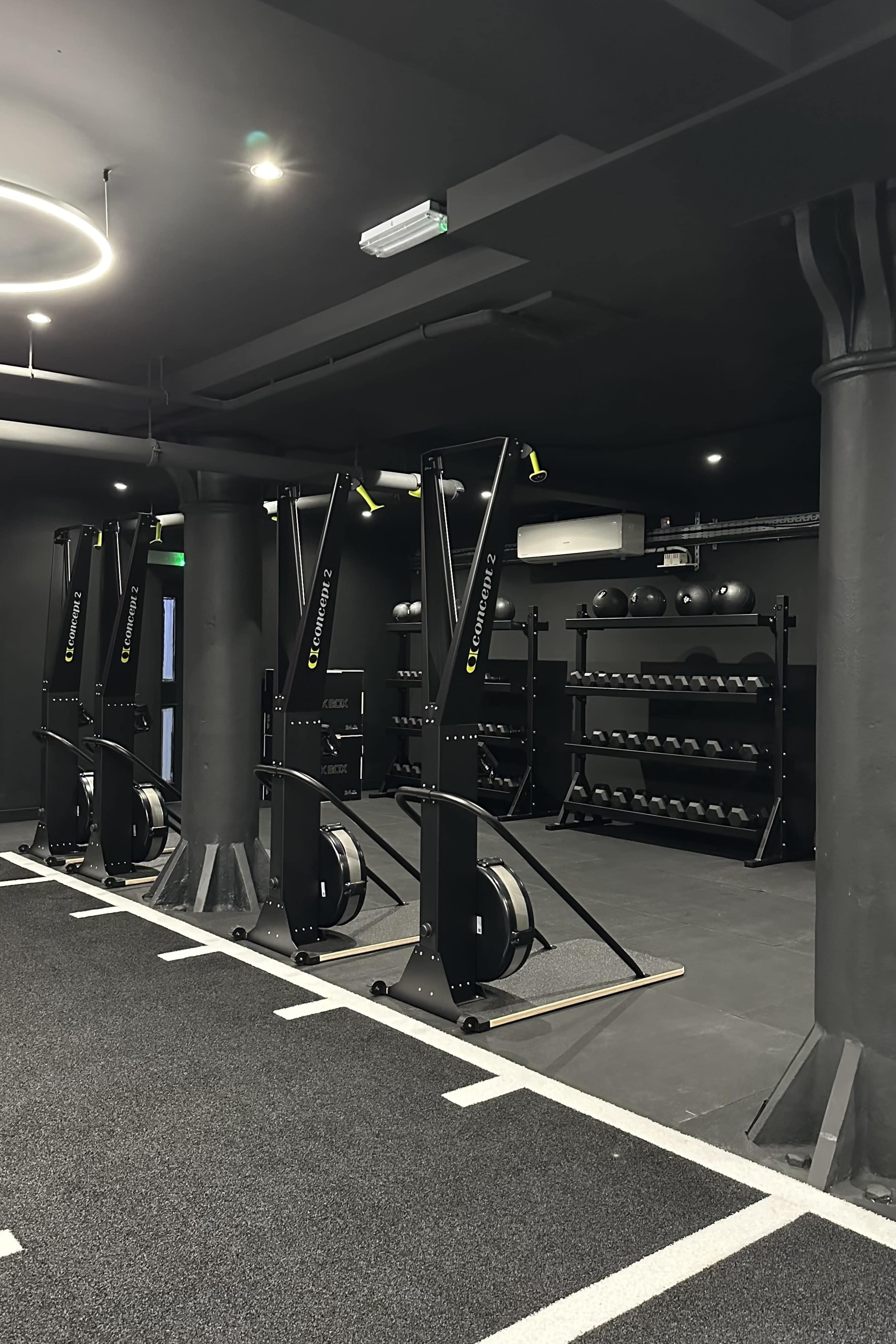 The image shows a modern gym interior featuring rowing machines and a rack of black weights on a dark, carpeted floor.