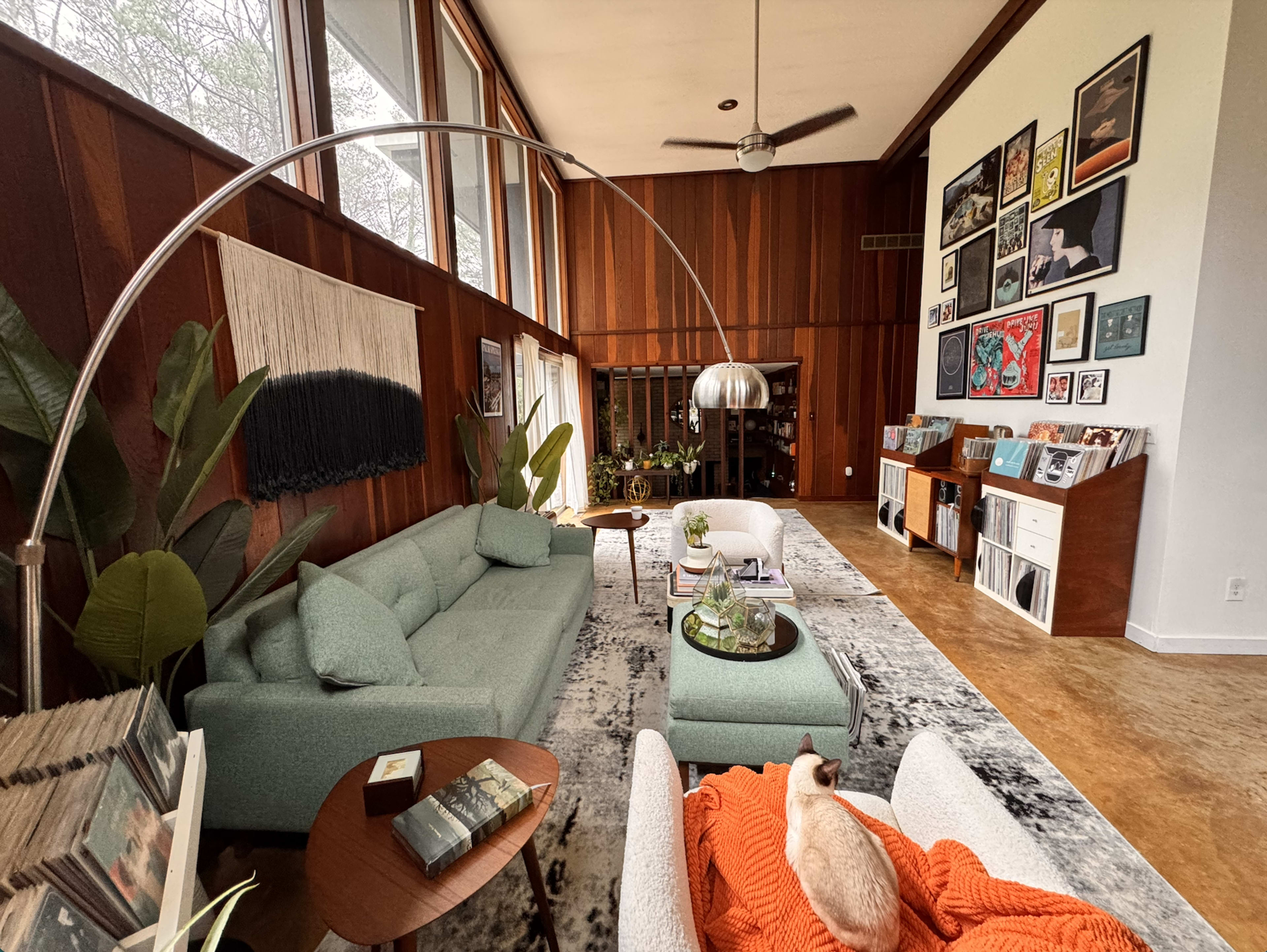 The image shows a spacious living room with large windows, a sectional sofa, a coffee table, and various artworks displayed on the walls.