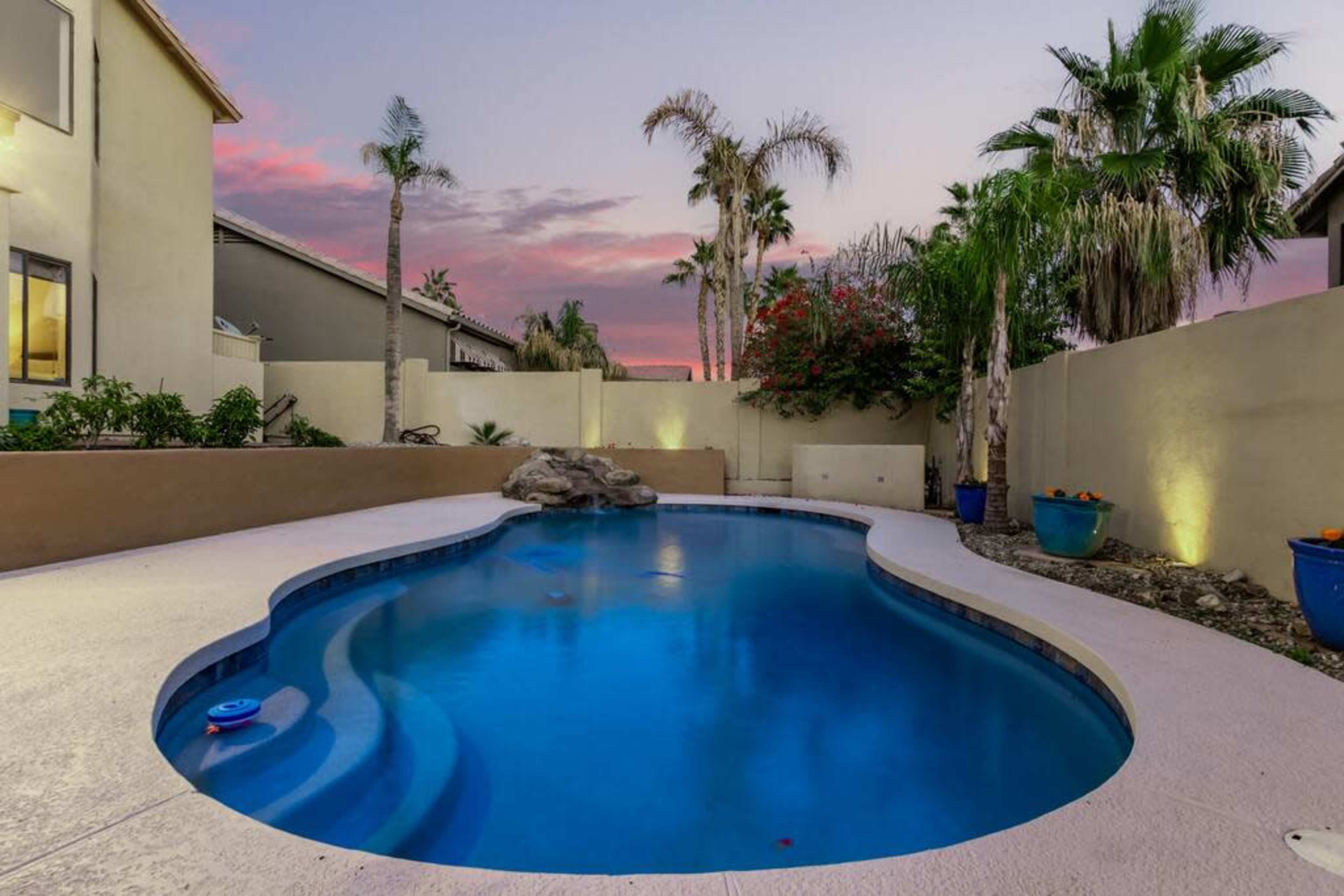 A backyard swimming pool surrounded by palm trees and colorful potted plants at dusk.