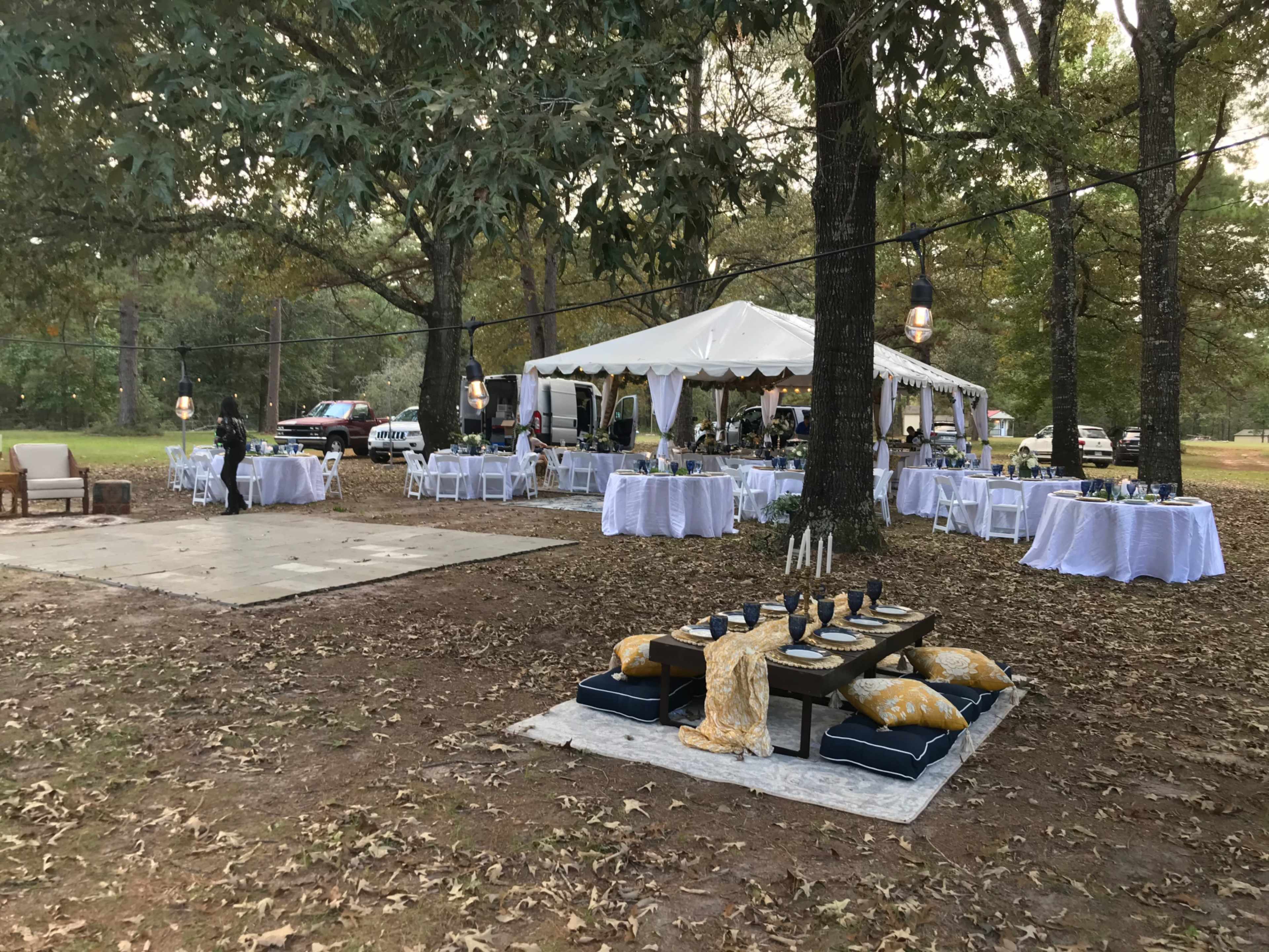 A large outdoor event setup features a tent with tables, chairs, and decorative elements, surrounded by trees and a grassy area.