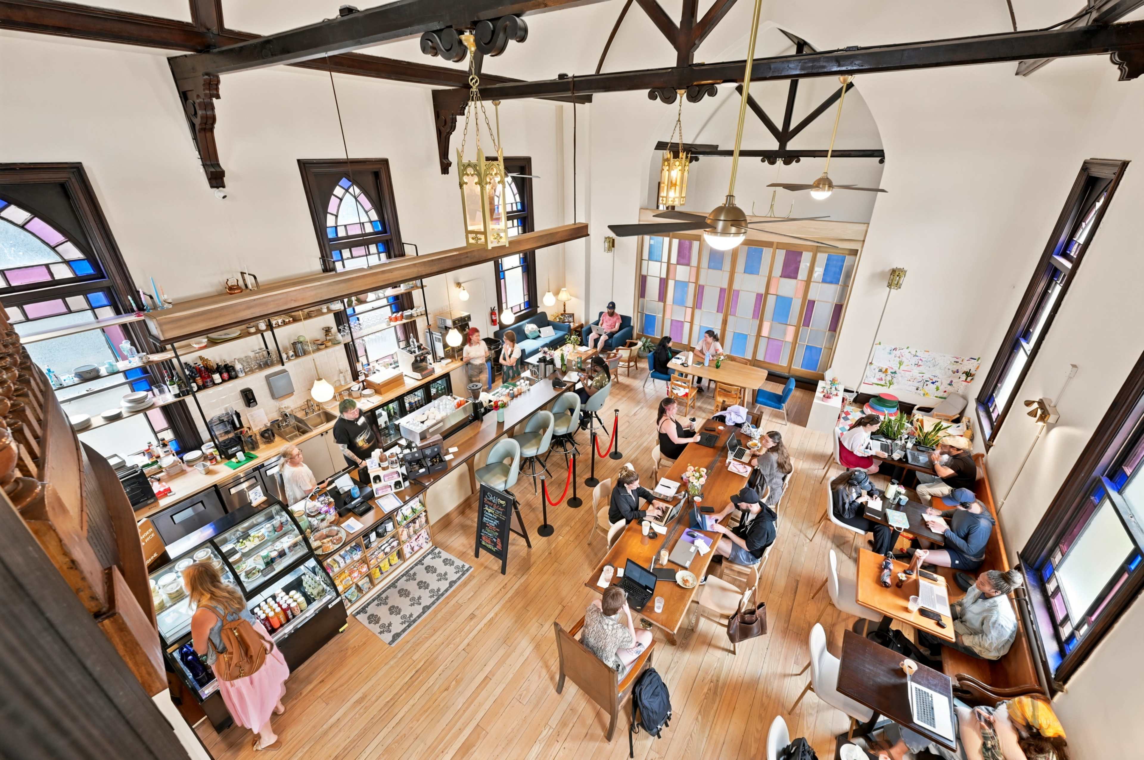 The image shows a busy café with customers seated at various tables, a counter displaying food and drinks, and large windows allowing natural light to fill the space.