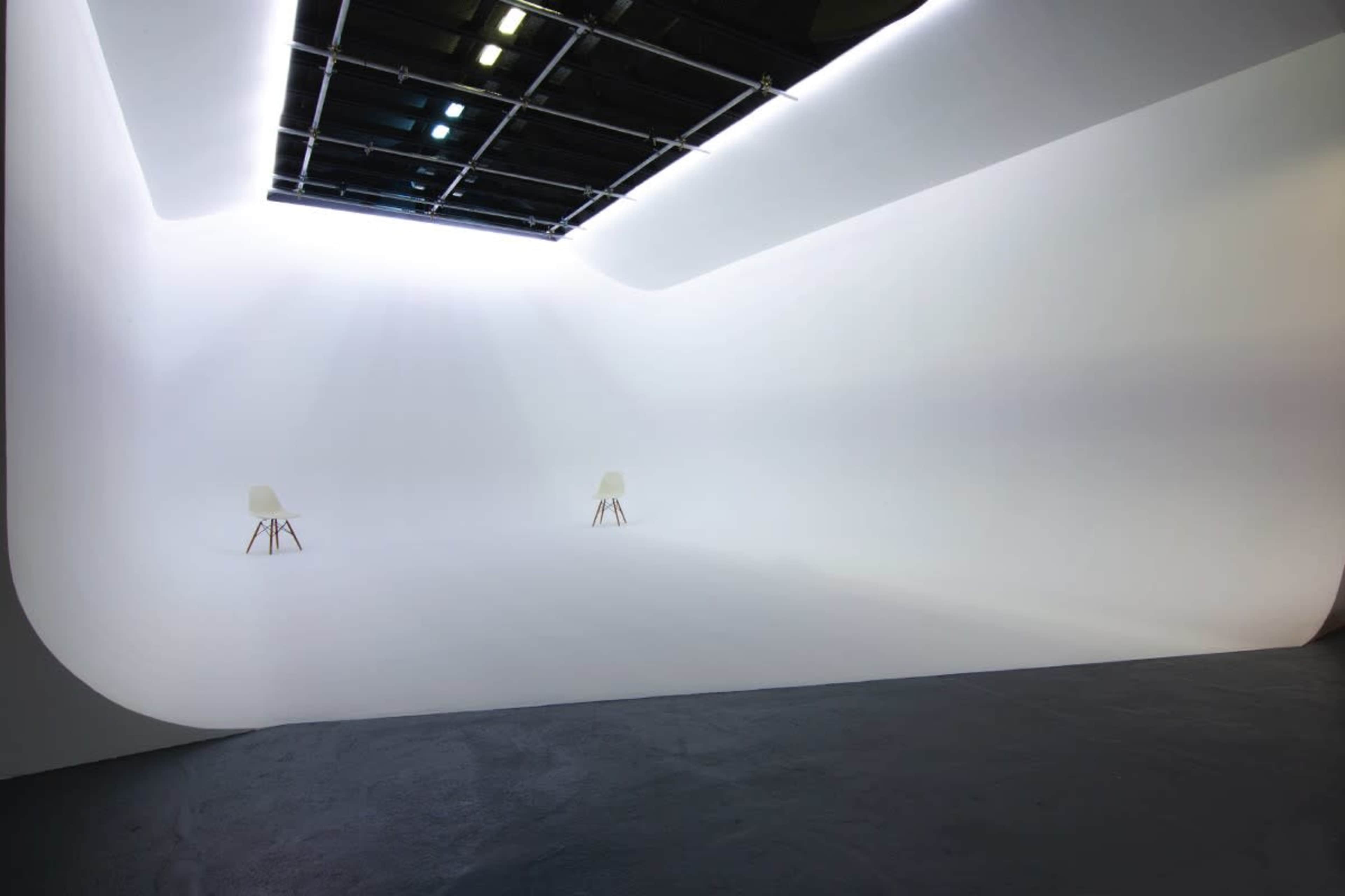 The image shows a spacious, empty studio with a seamless white background and two chairs set apart in the center.