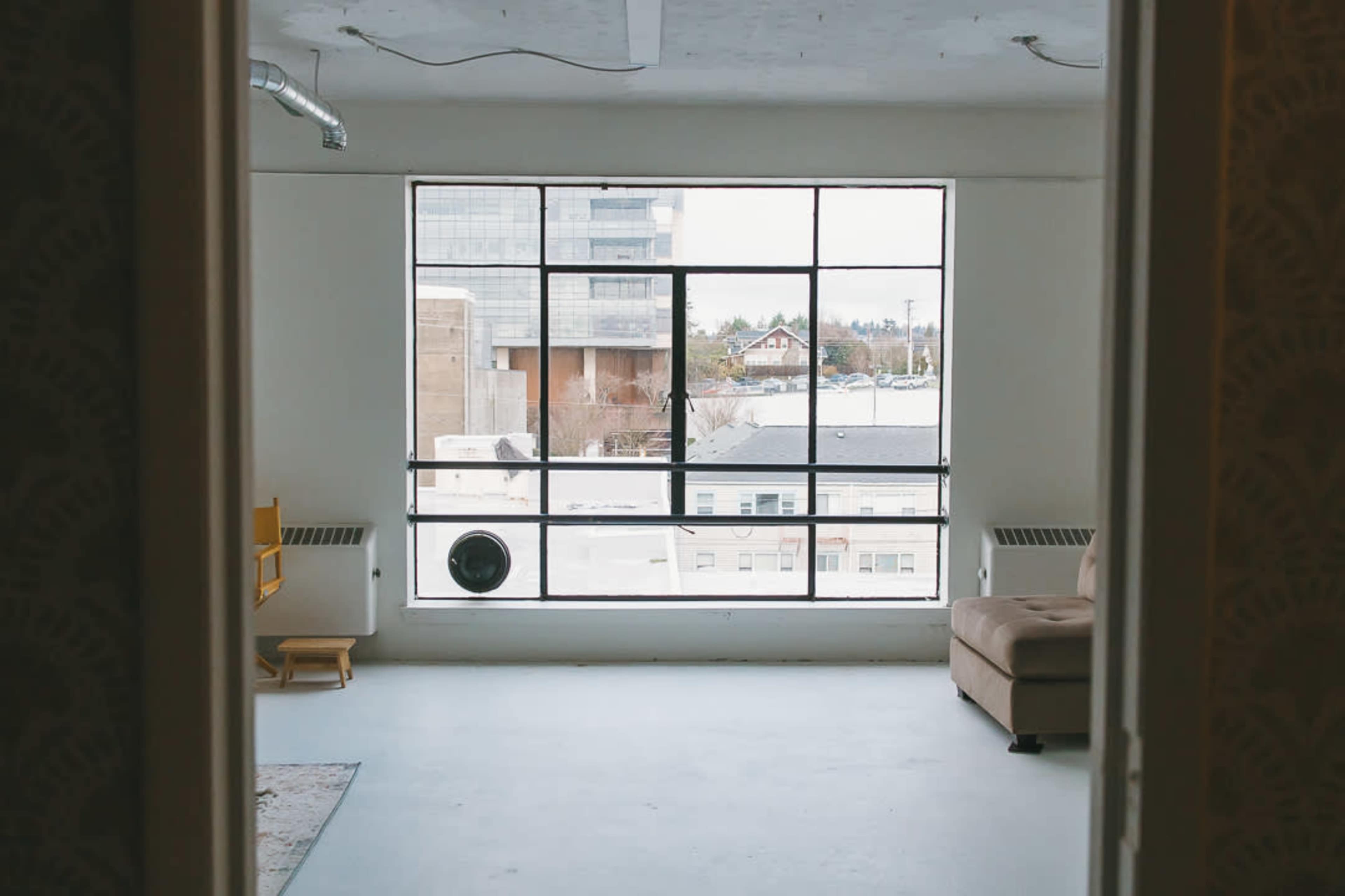 The image shows a spacious room with a large window displaying a cityscape view, featuring a couch and a small chair in the foreground.