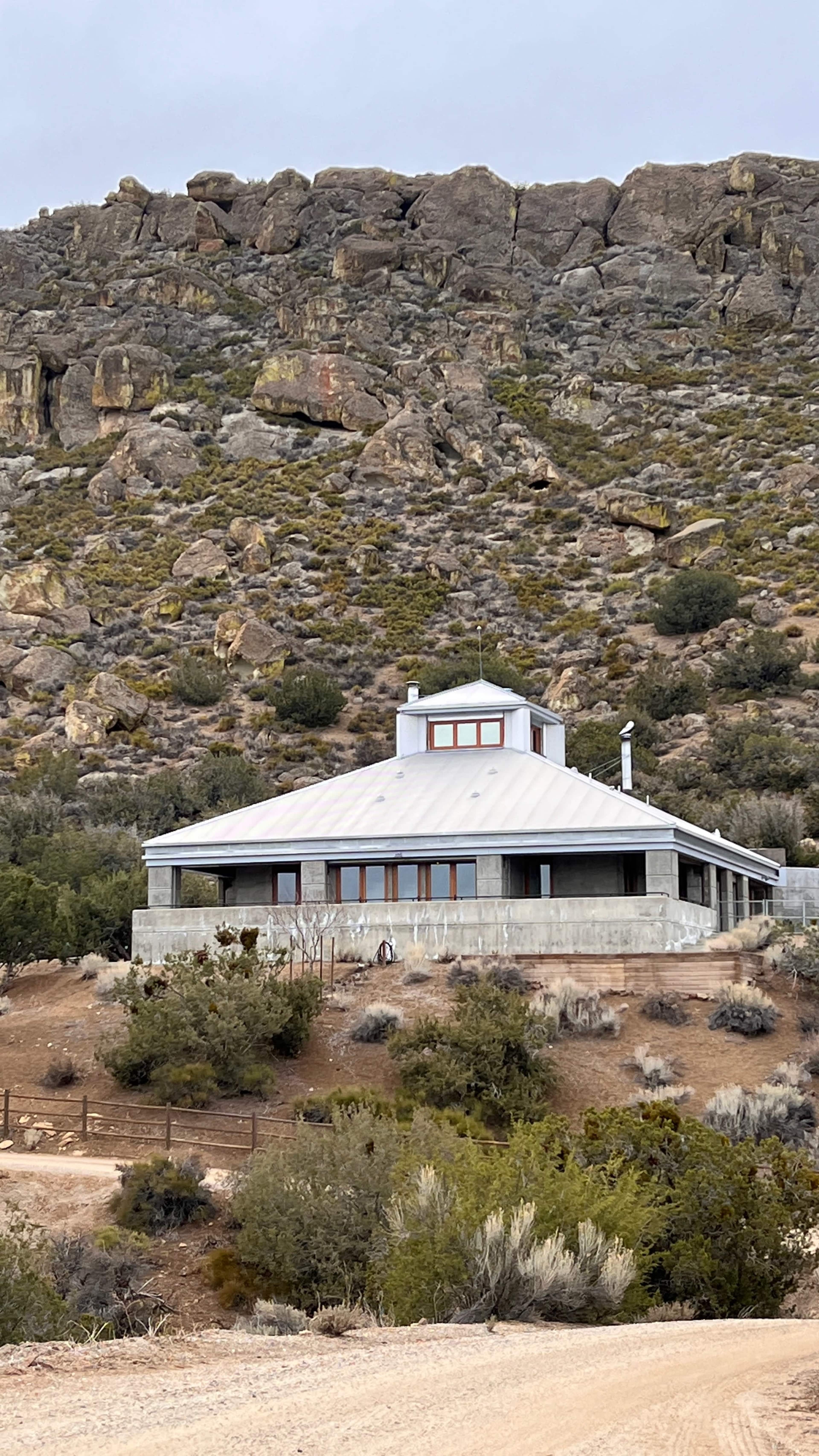 A modern, circular house with large windows is situated on a hillside, surrounded by sparse vegetation and rocky terrain.