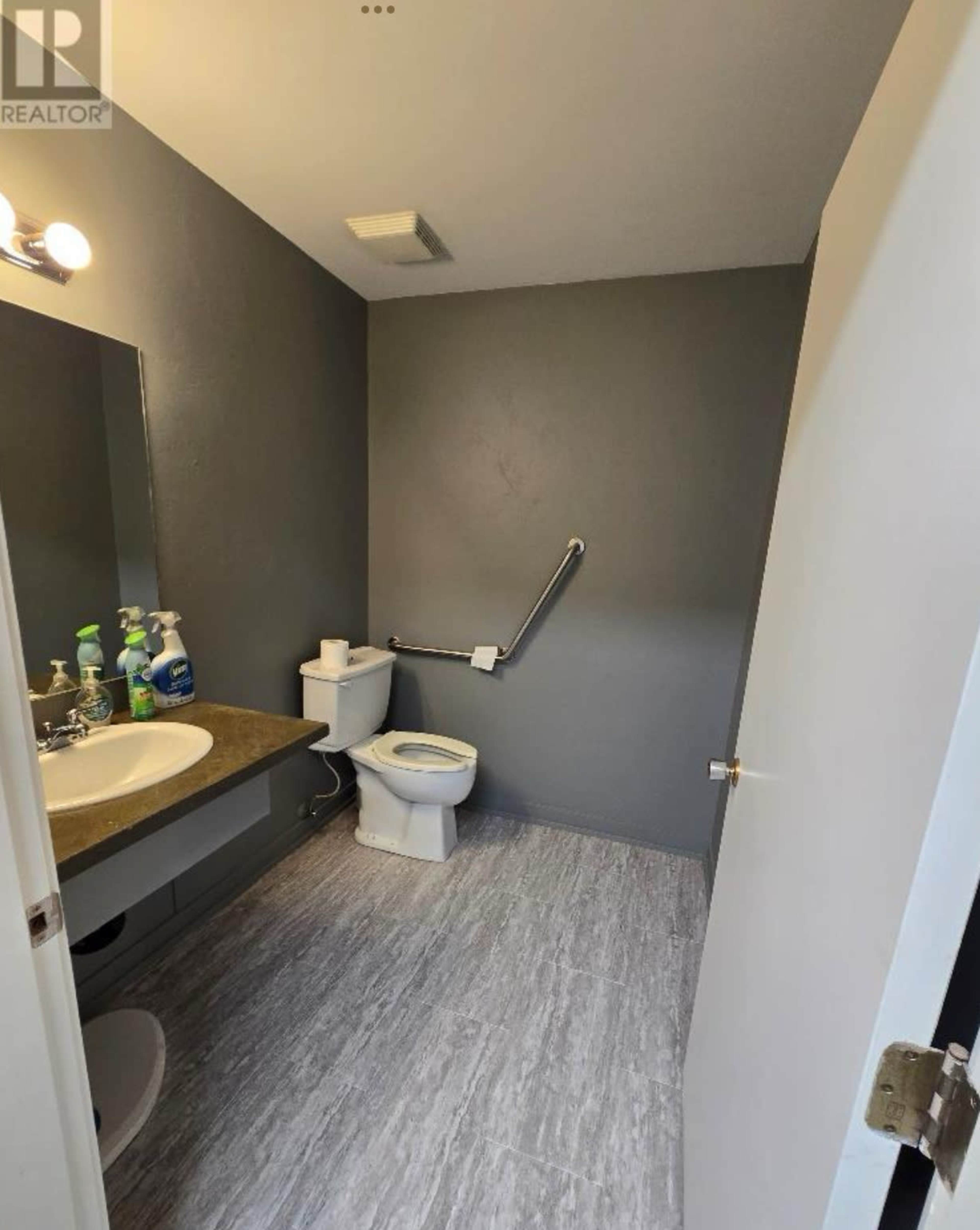 The image shows a bathroom with a sink, toilet, grab bar, and a mirror, featuring grey walls and a light-colored floor.