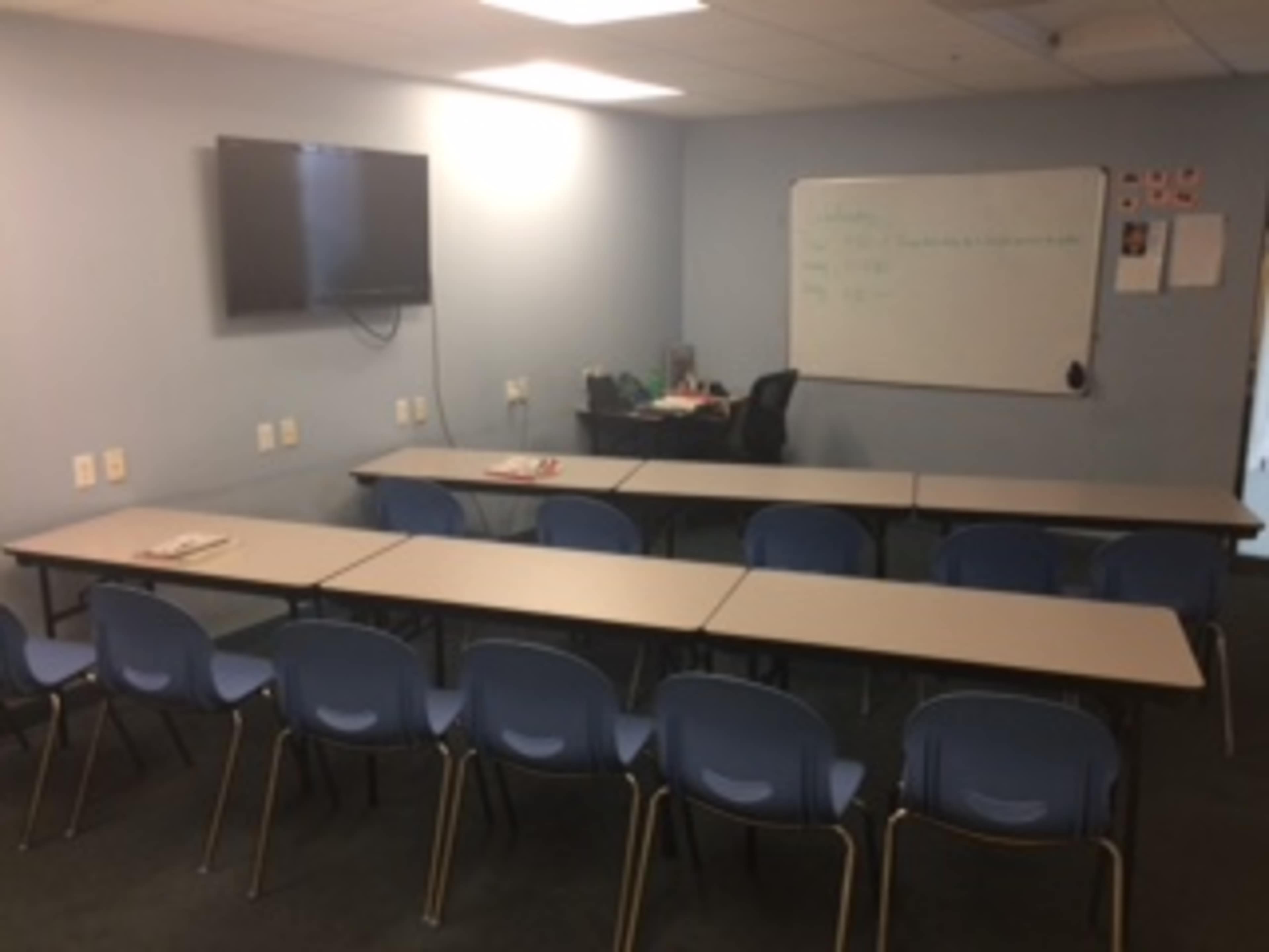 A classroom with several rows of tables and chairs, a whiteboard, and a television mounted on the wall.