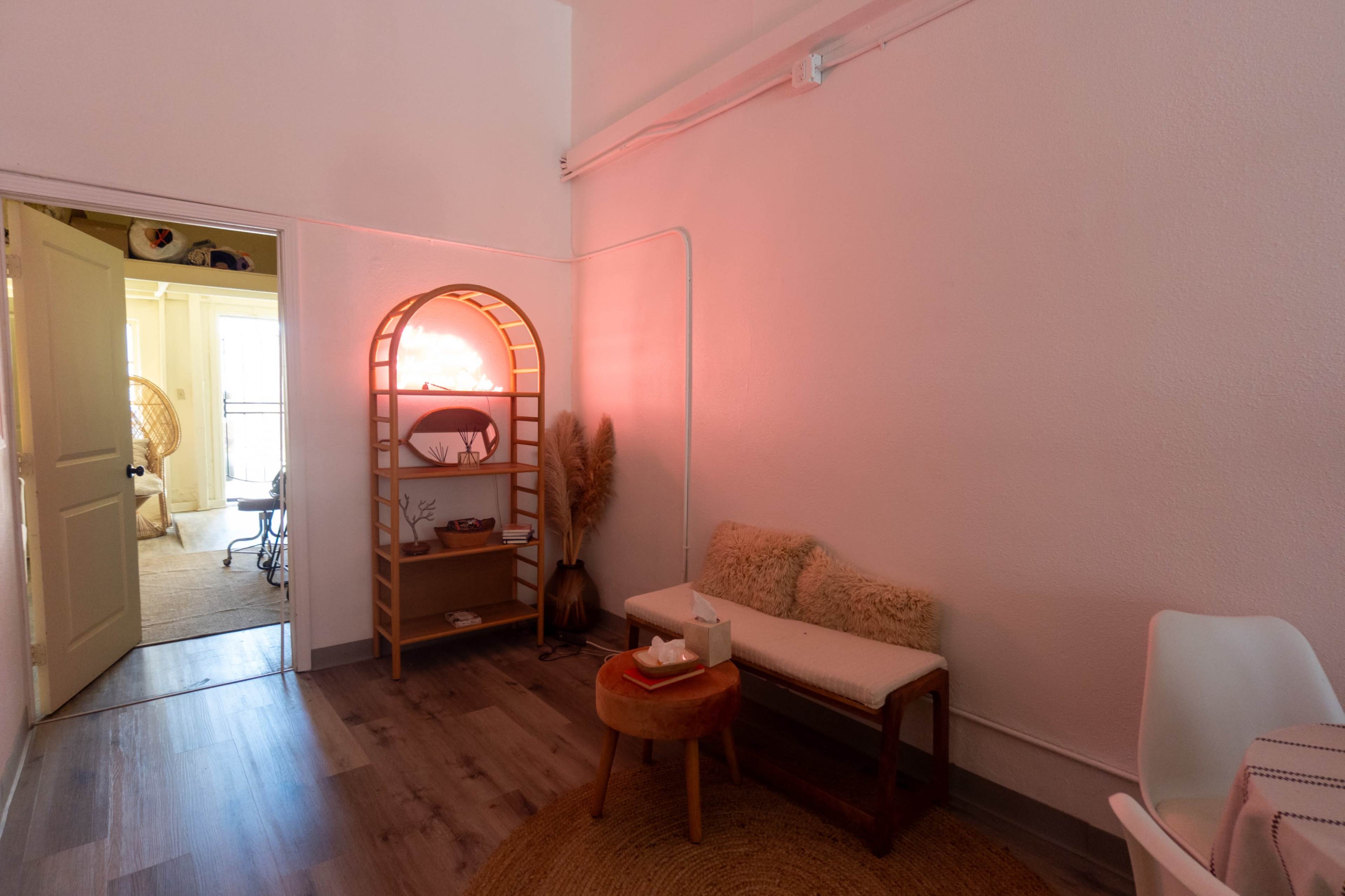 A softly lit interior space features a small seating area with a bench and a round table, alongside a shelf displaying decorative items.