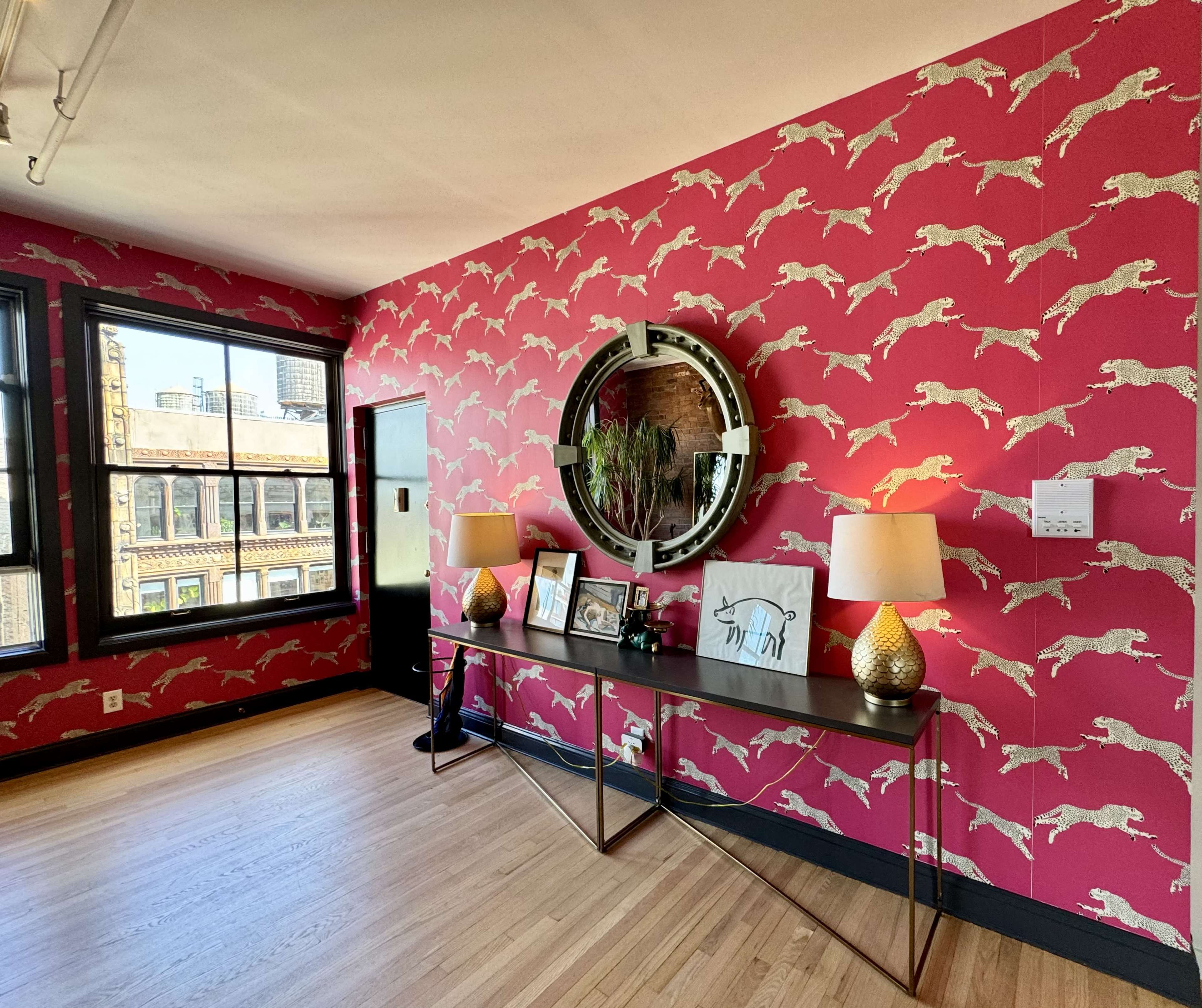 The image shows a room with a vibrant pink wall featuring a pattern of leaping dogs, a circular mirror, a console table with decorative items, and two lamps.