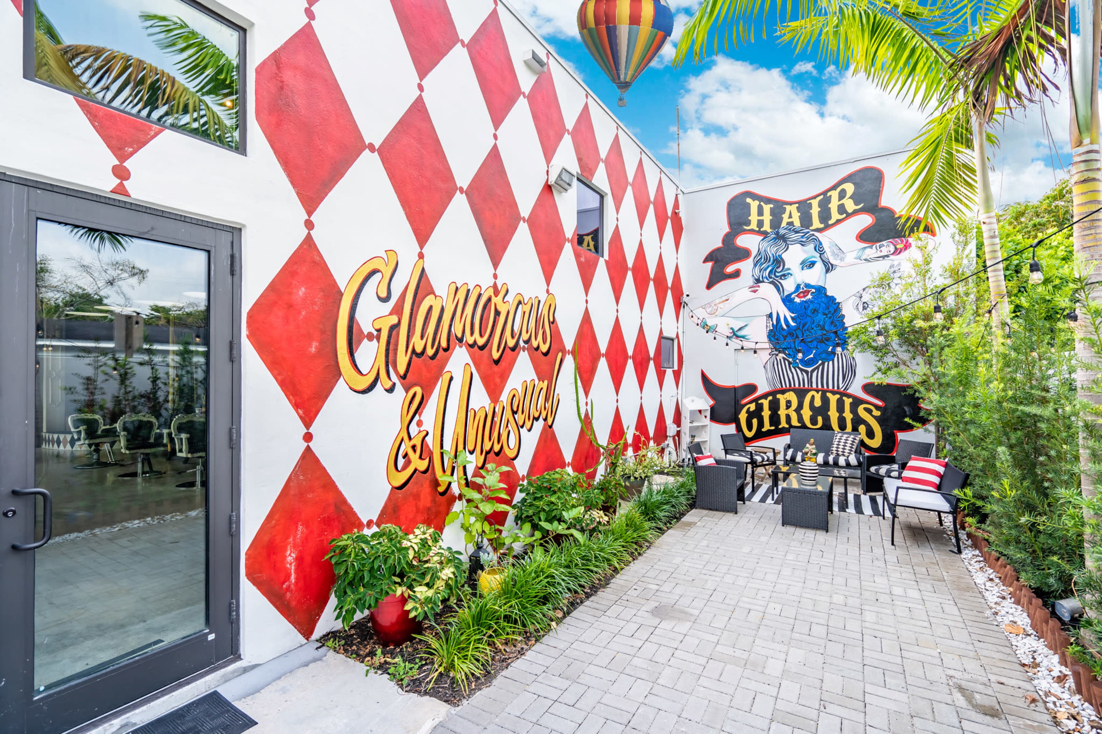 A colorful outdoor space with a checkered red and white wall featuring the text "Glamorous & Amused" and a large mural of a woman with the words "Hair Circus" nearby, surrounded by plants and seating.