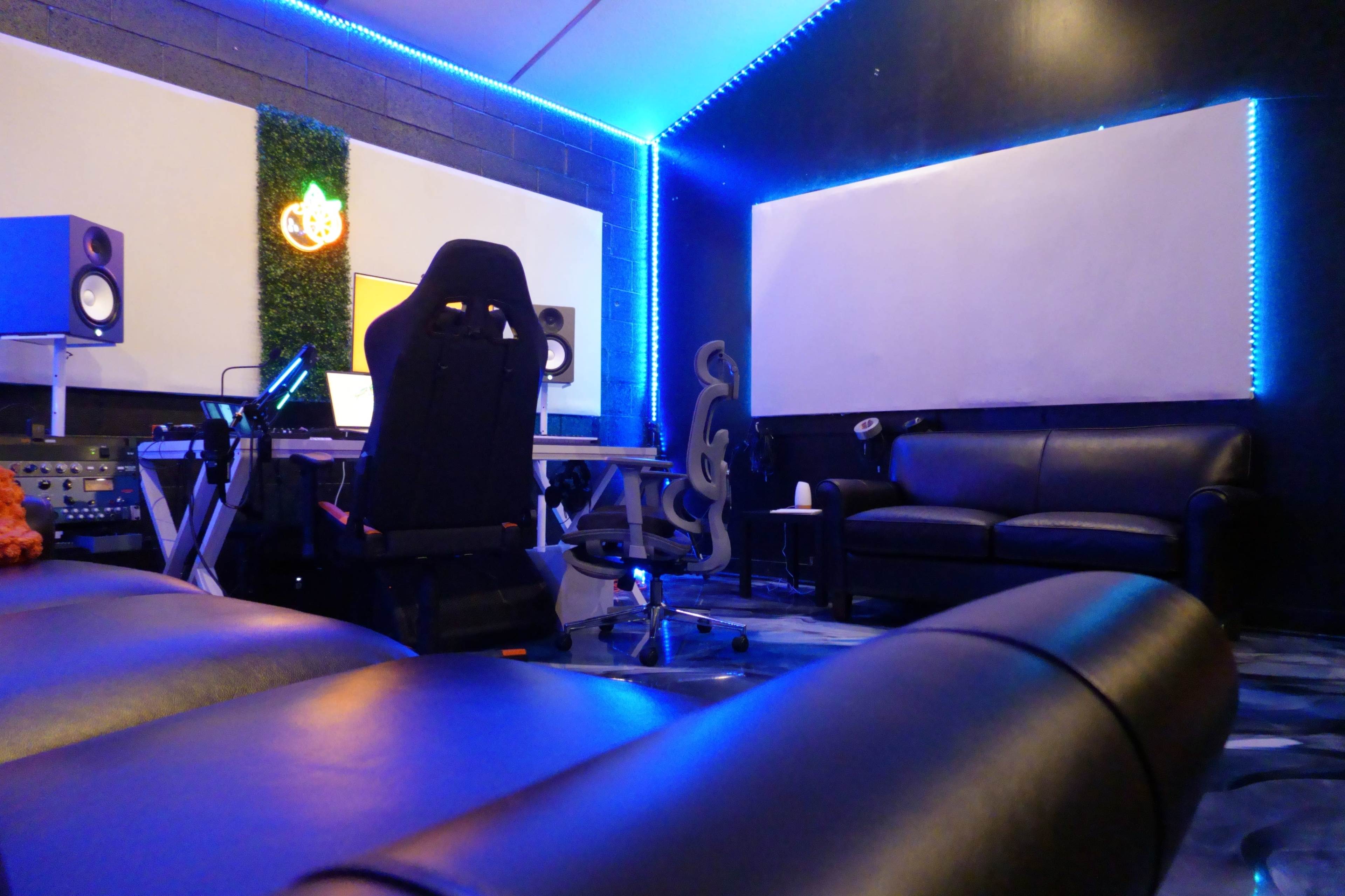 The image shows a music production studio with a black leather couch, a desk with equipment, and blue LED lighting around the walls.