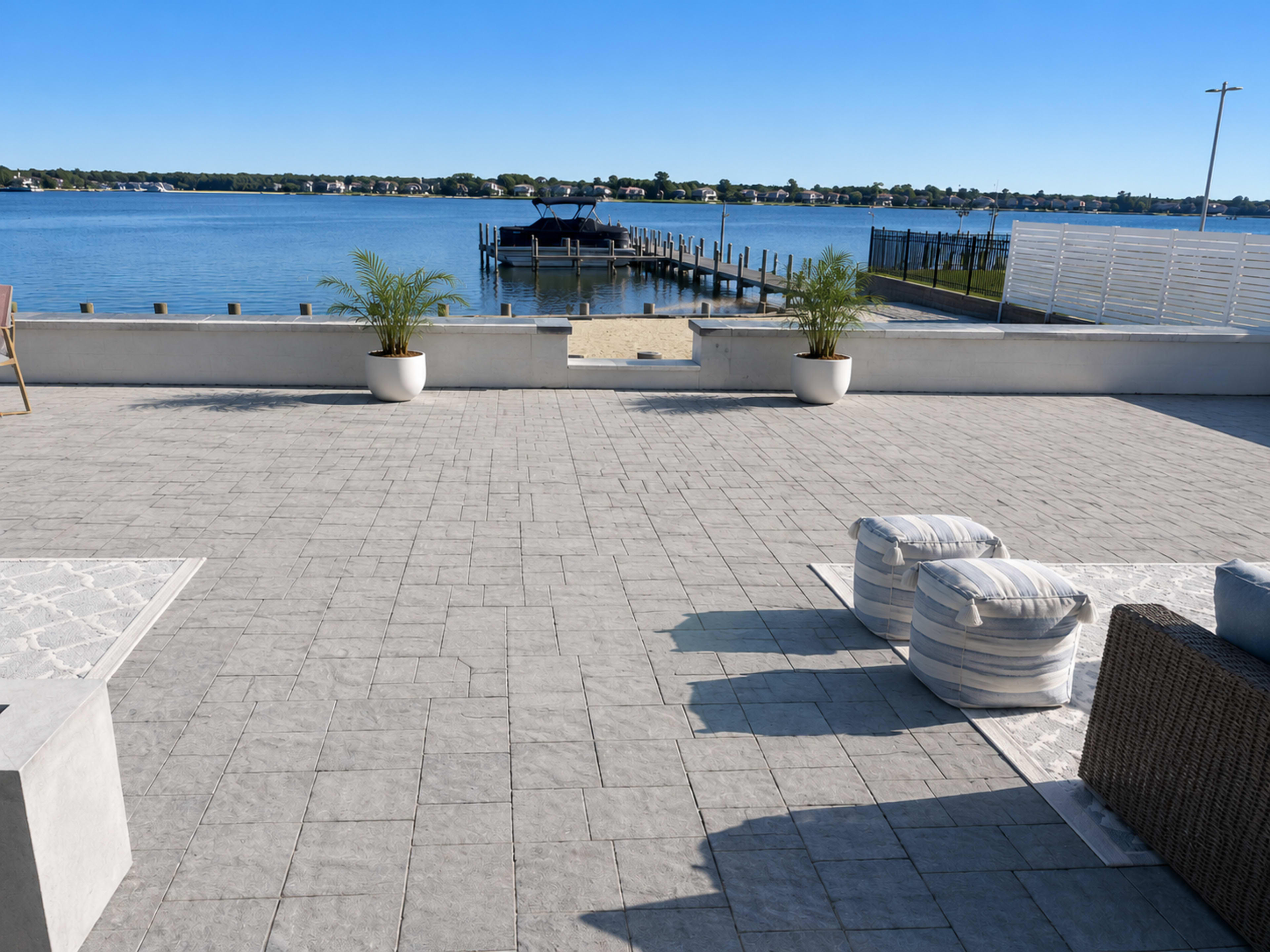 The image shows a large patio area with a view of a calm body of water, a dock, and boats in the distance.