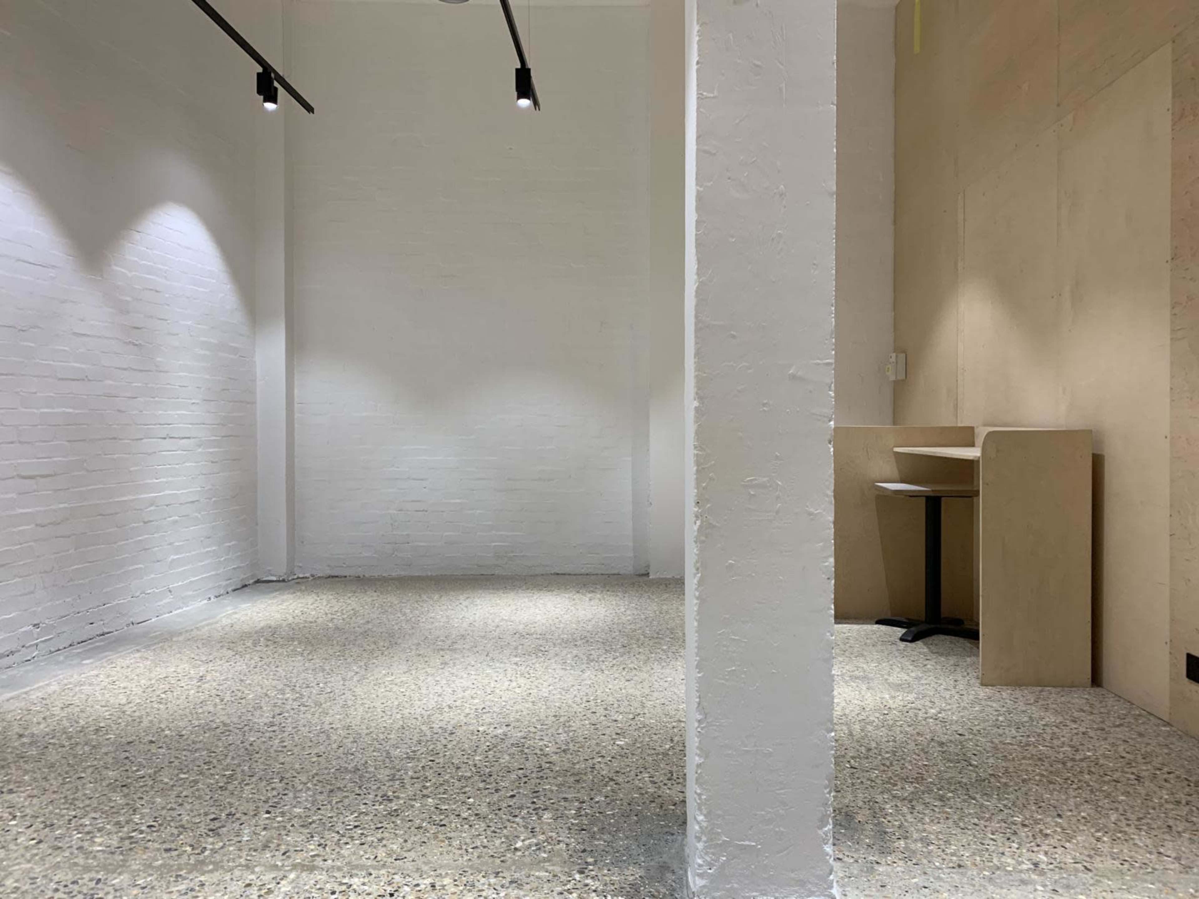 A sparse, well-lit interior space with a light-colored wall, a concrete floor, and a solitary table and chair positioned against one side.