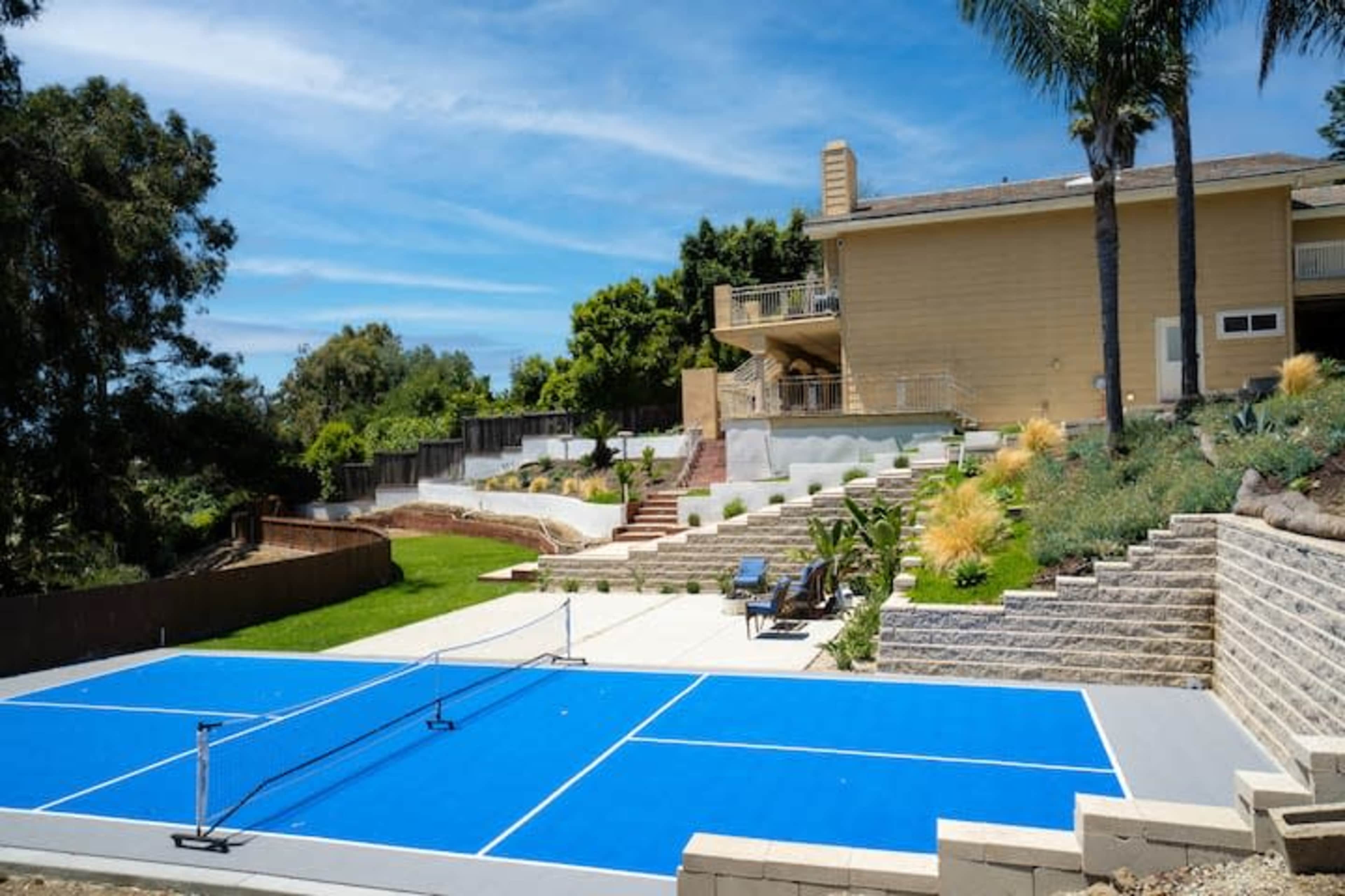 A blue tennis court is situated in a landscaped backyard in front of a two-story house.