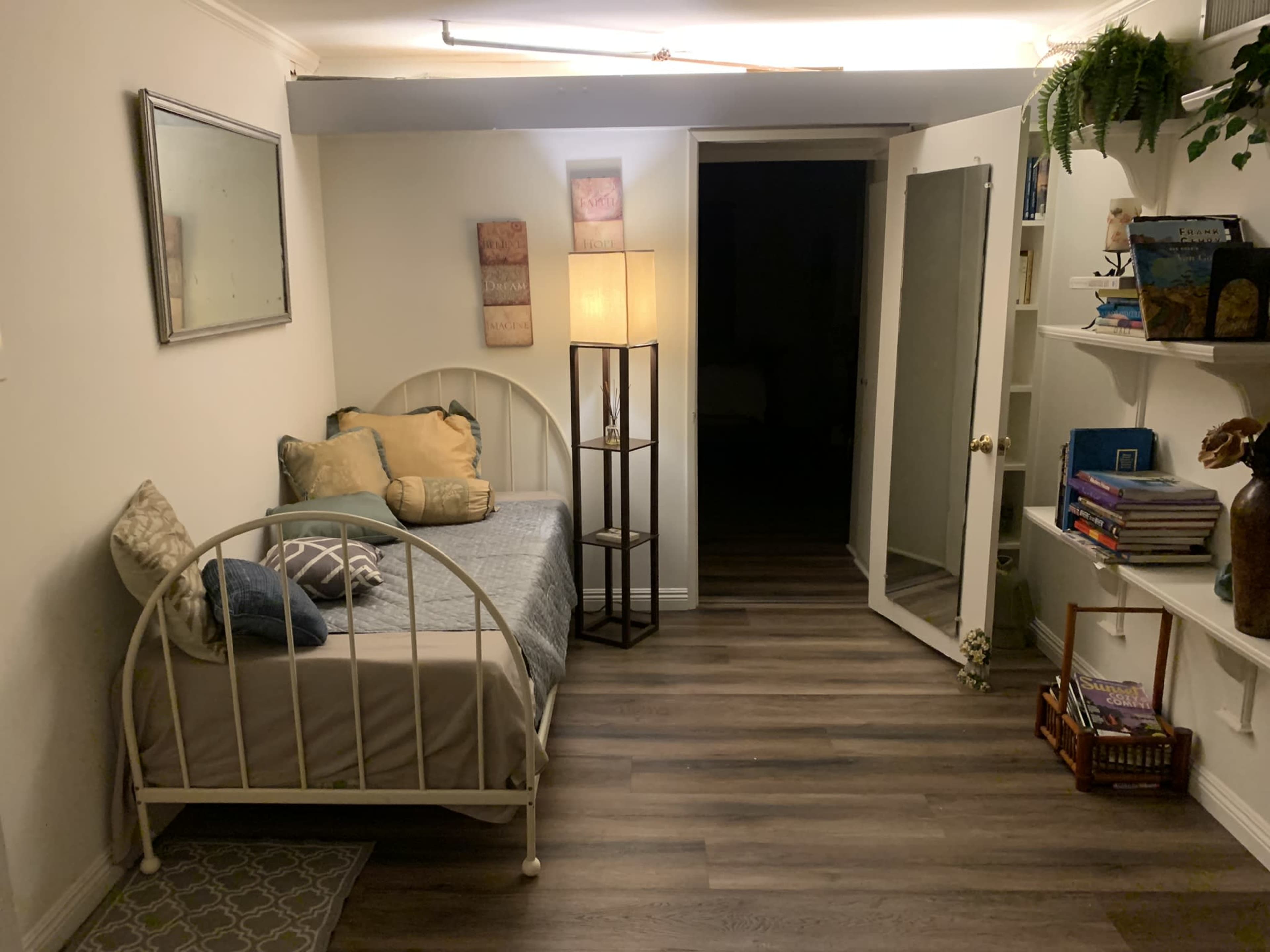 A small room with a bed against one wall, a lamp beside it, and a mirror next to an open door leading to a darker space.