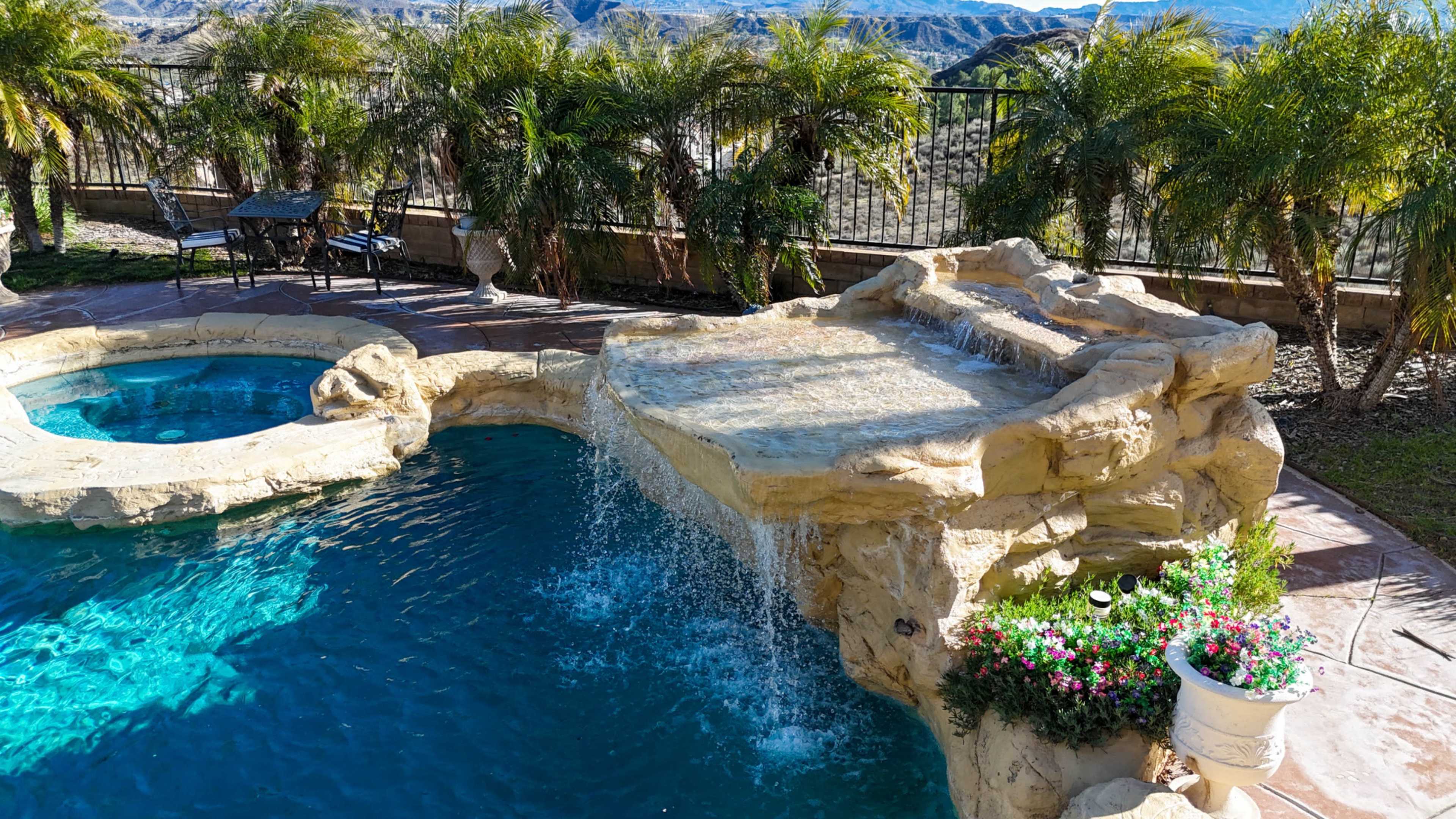 A pool area features a rock waterfall cascading into the water, surrounded by palm trees and a stone patio with a table and chairs.