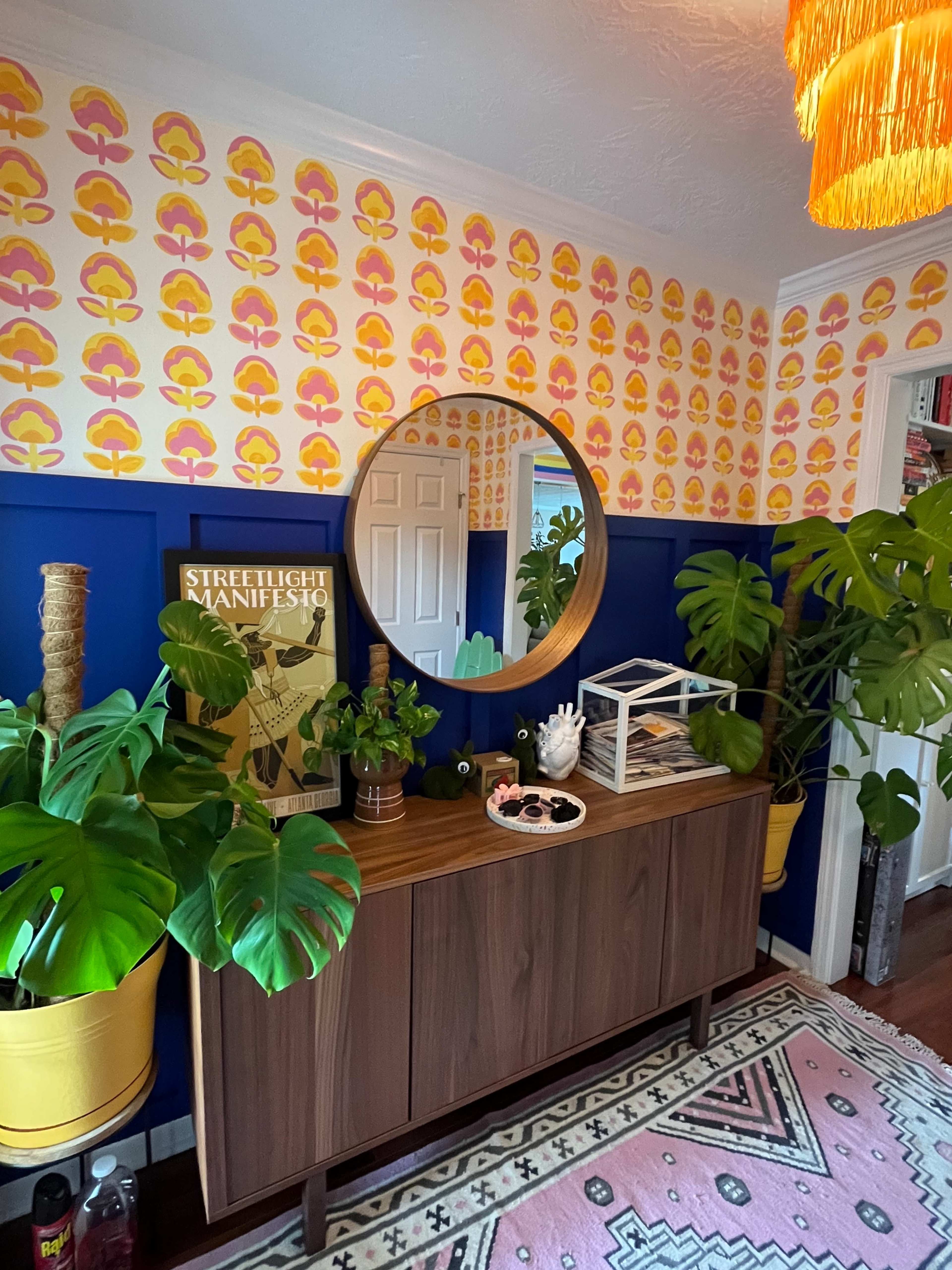 The image features a living room corner with a sideboard, circular mirror, colorful wallpaper, and various plants, creating a vibrant interior space.