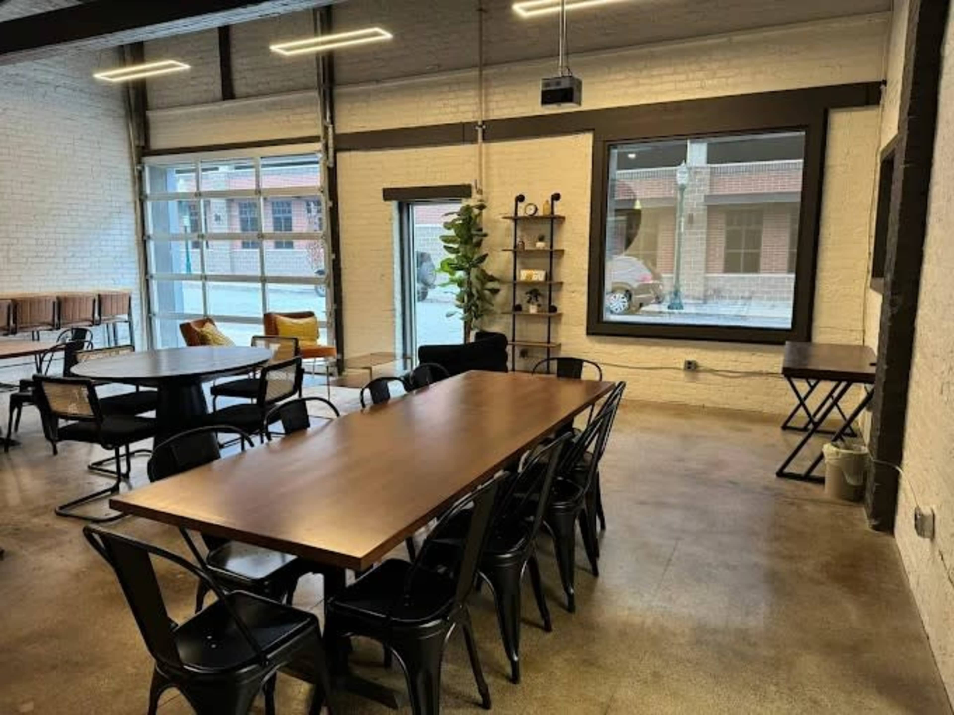 The image shows a modern open space featuring a large wooden table surrounded by black chairs, with large windows letting in natural light and a few pieces of furniture against the walls.