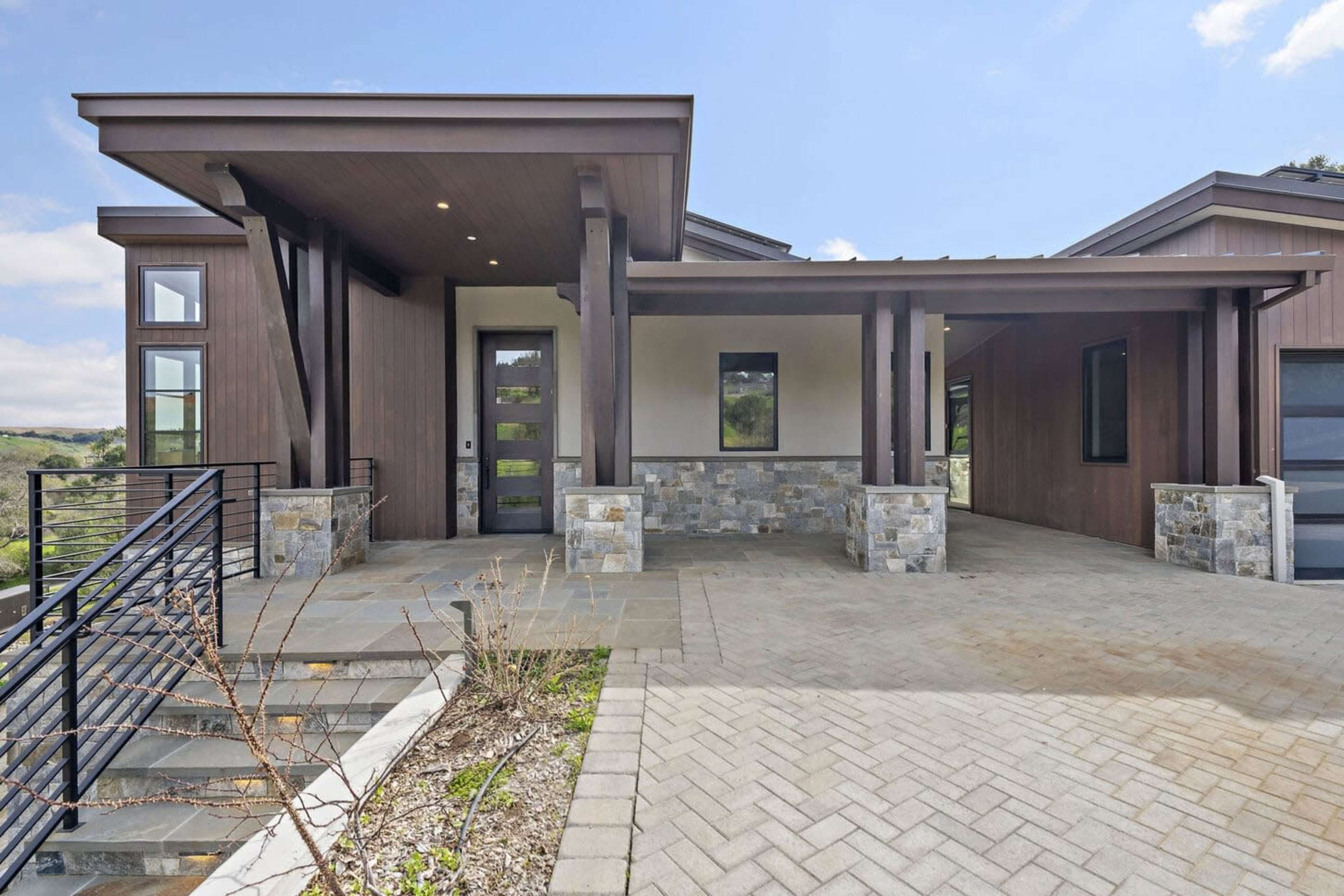 The image shows a modern house with a combination of wooden and stone exterior, featuring a covered entrance and a landscaped front area.