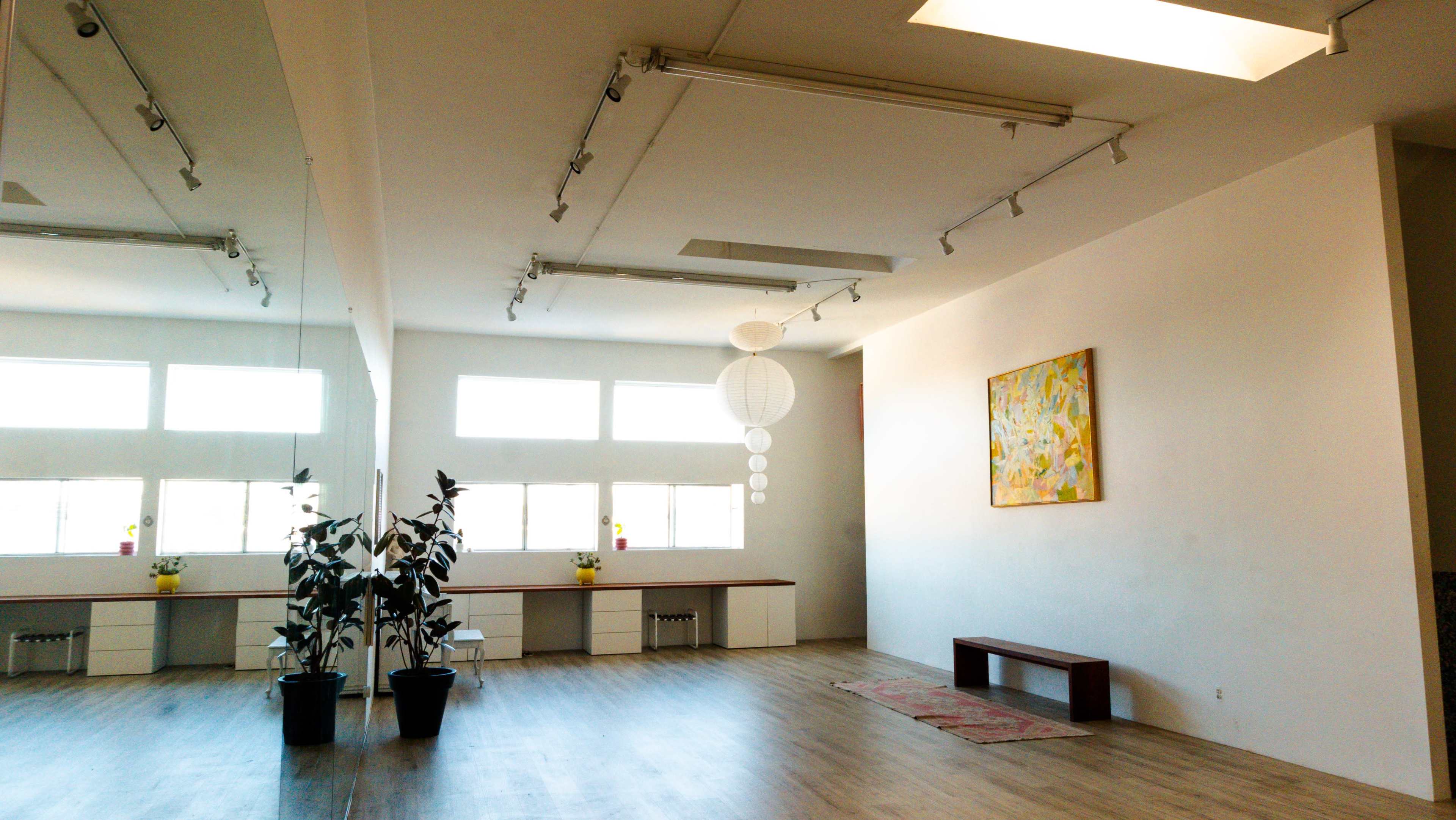 The image shows a bright, minimalistic room with large windows, wooden flooring, a simple bench, a painting on the wall, and a potted plant.